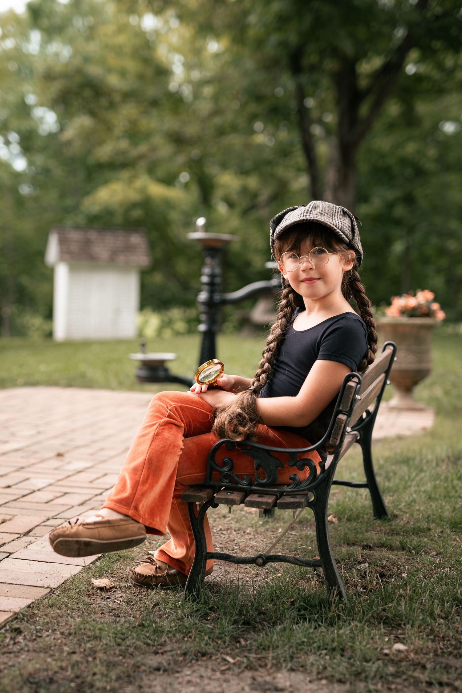 Smiling girl in orange pants holds magnifying glass while seated on decorative iron bench outdoors.