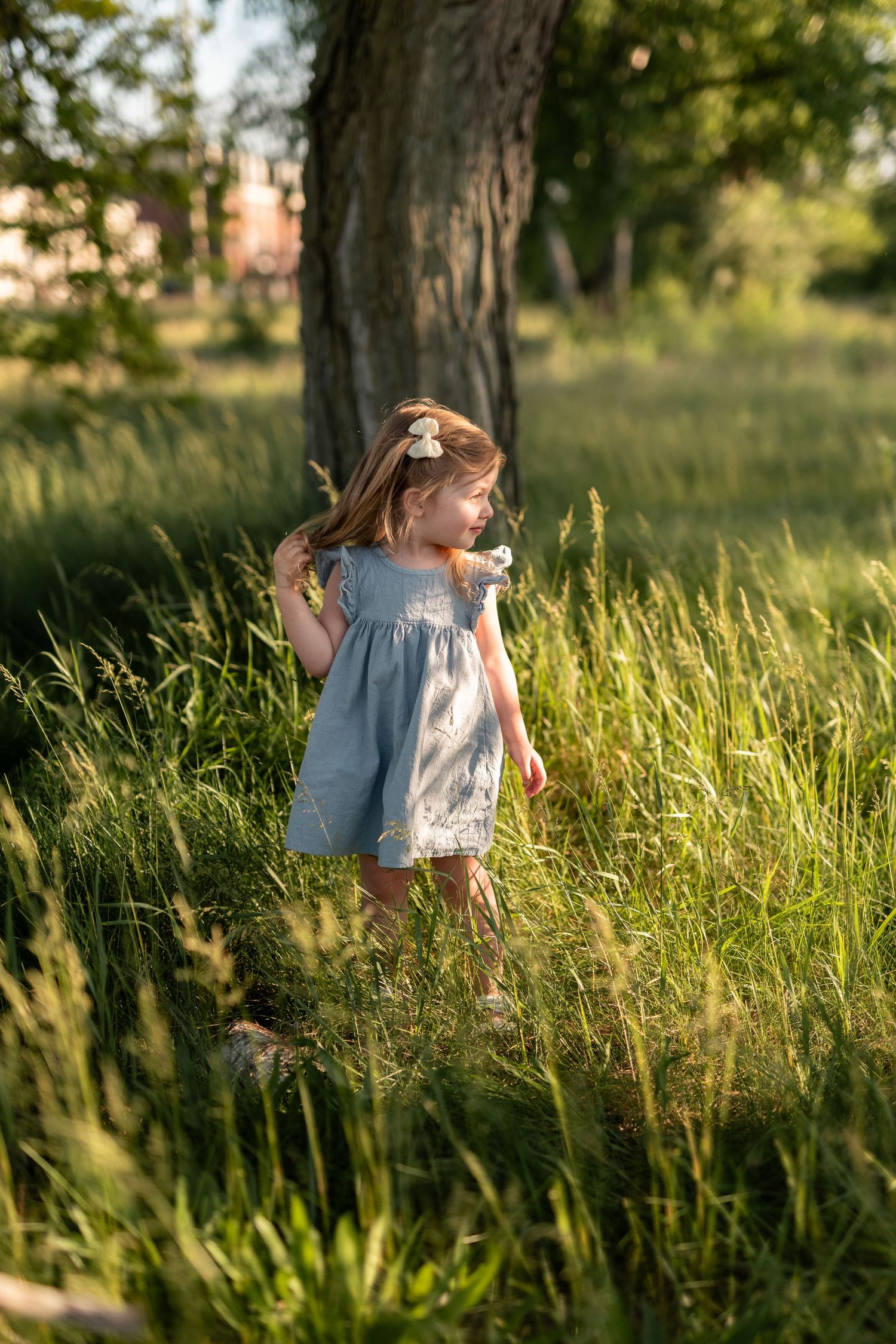 A young girl in a blue dress walks through tall green grass near a tree in golden hour sunlight.