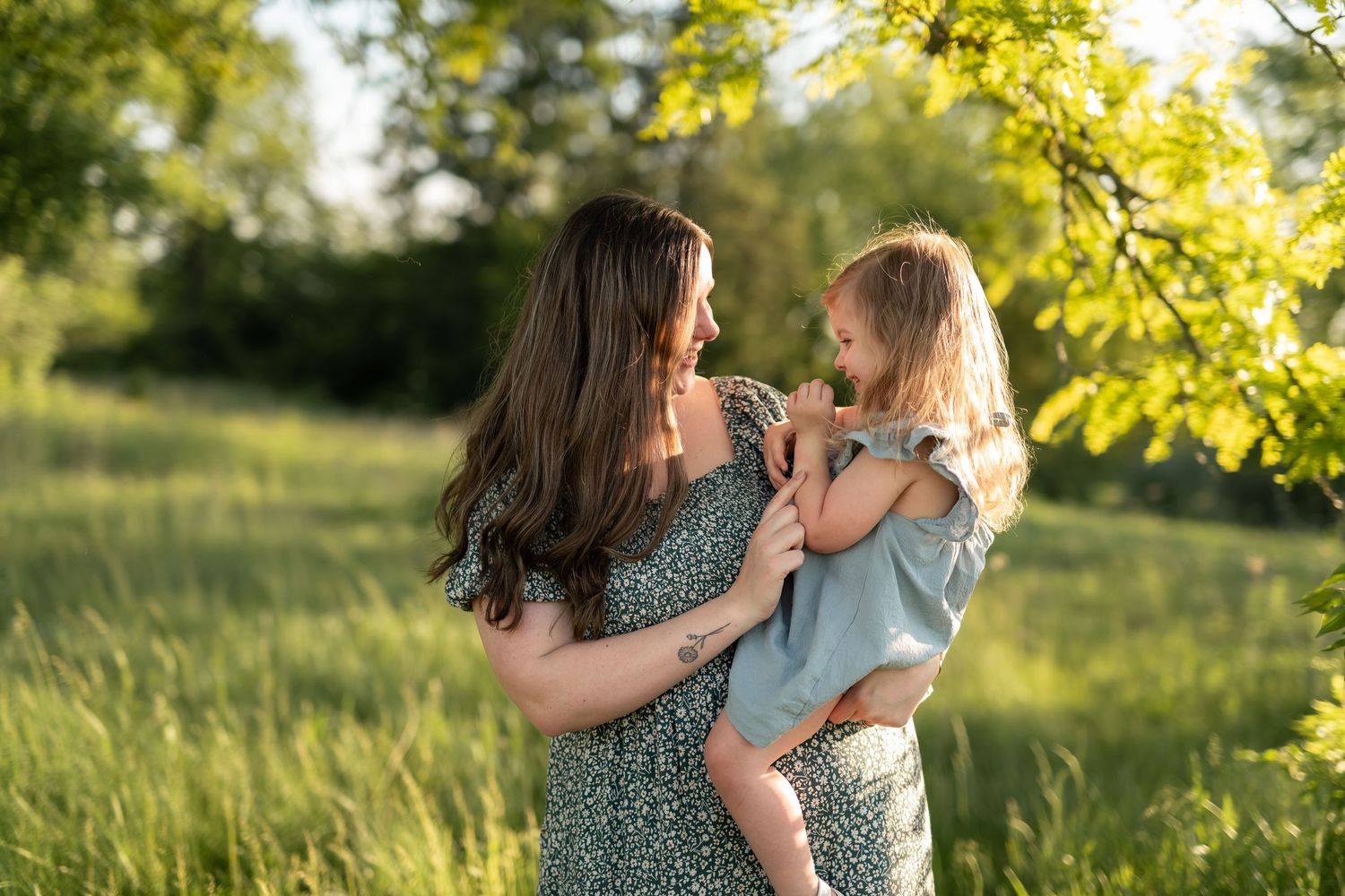 A mother holds her young daughter in a sunny green meadow, both smiling warmly in matching sage outfits.