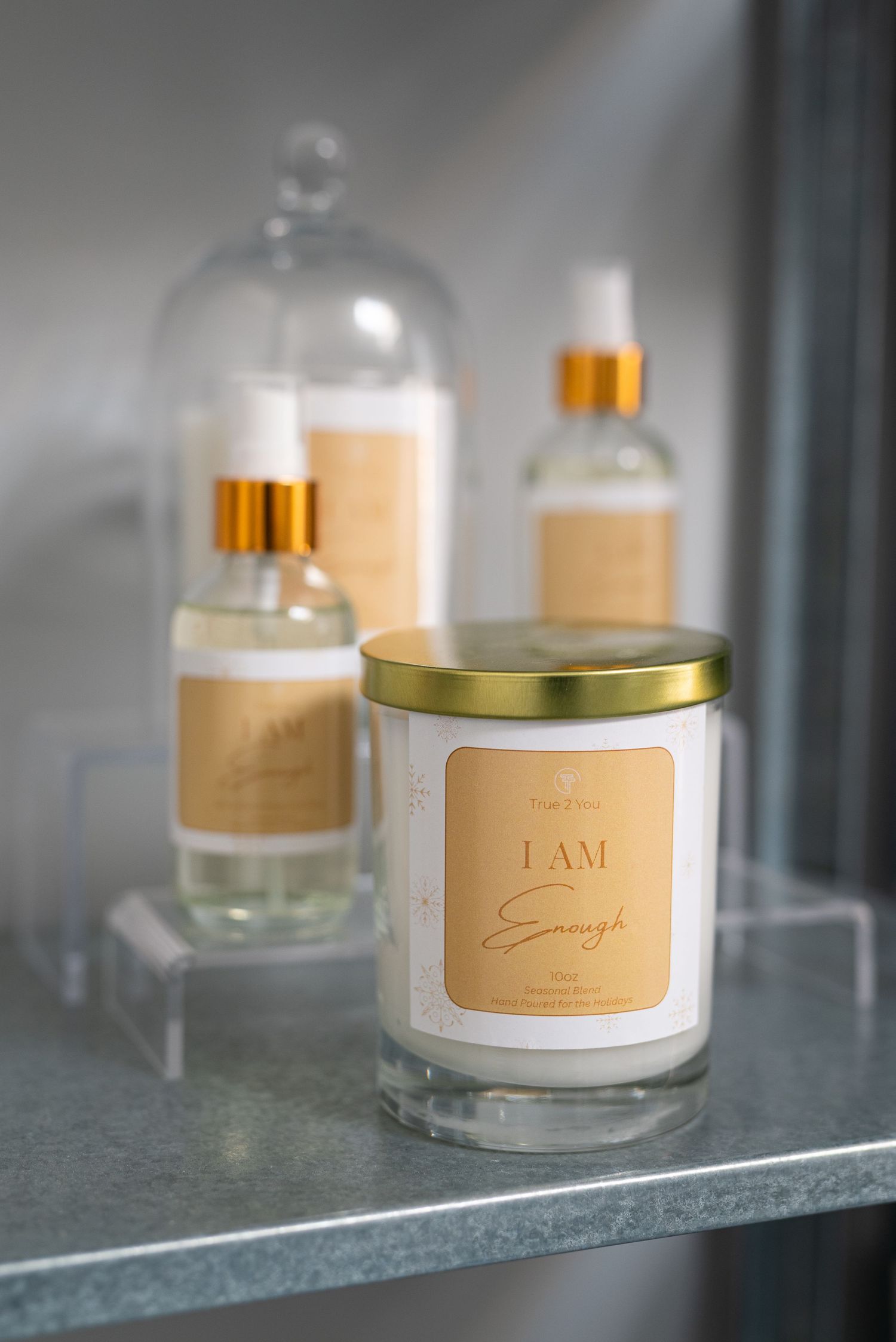 Glass candle and spray bottles with gold accents labeled 'I AM Enough' displayed on a shelf.