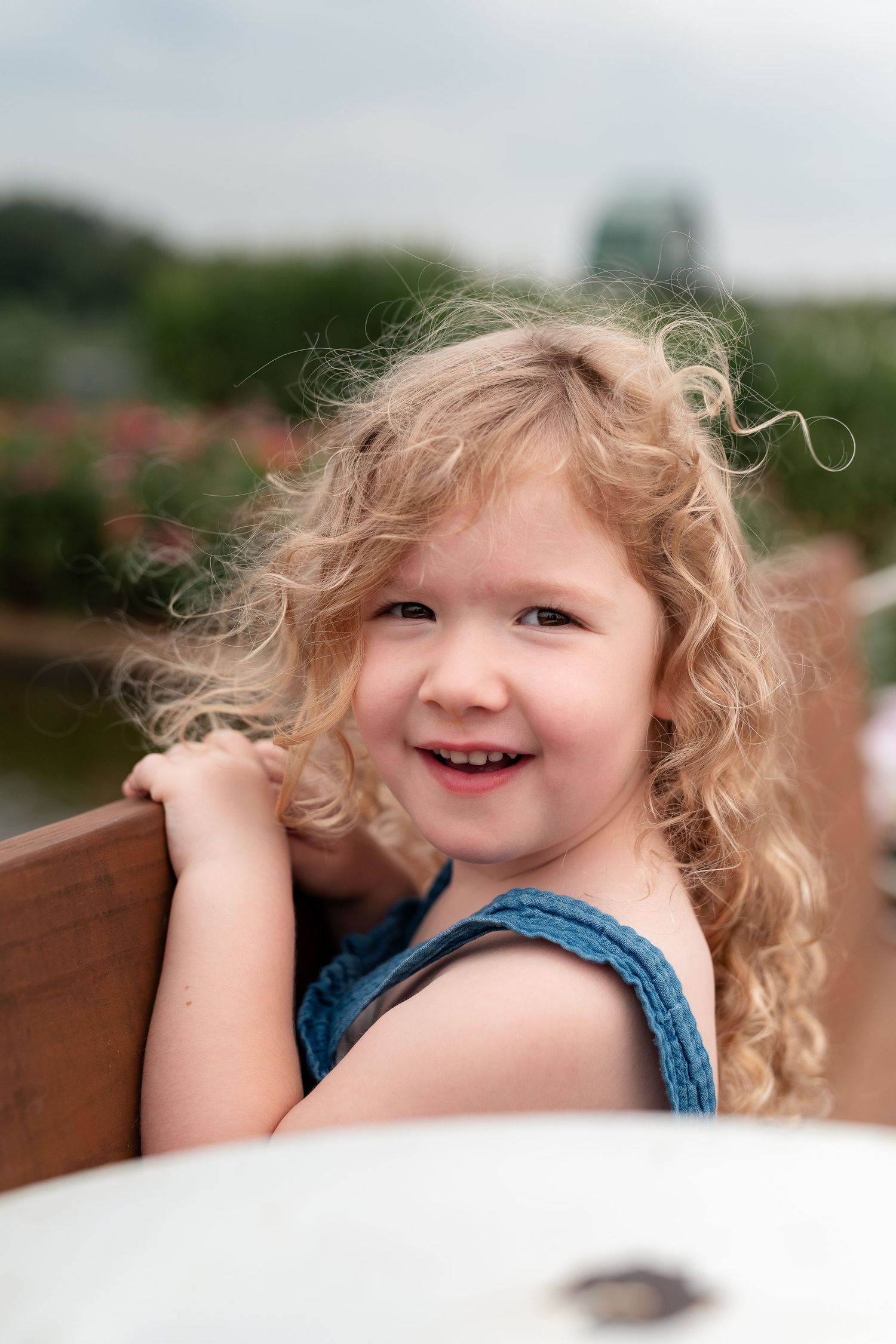 A cheerful curly blonde toddler girl in a blue outfit smiling brightly outdoors near a wooden fence.