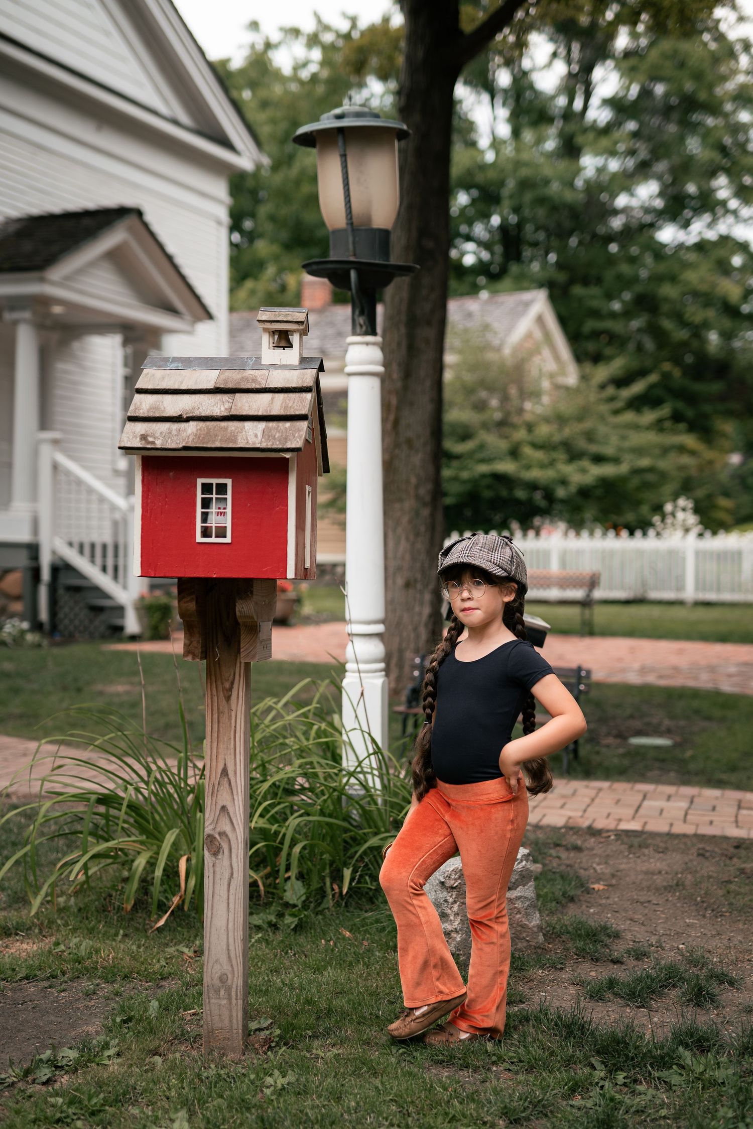 Young girl in orange pants and cap poses by a red birdhouse and lamp post in a suburban yard.