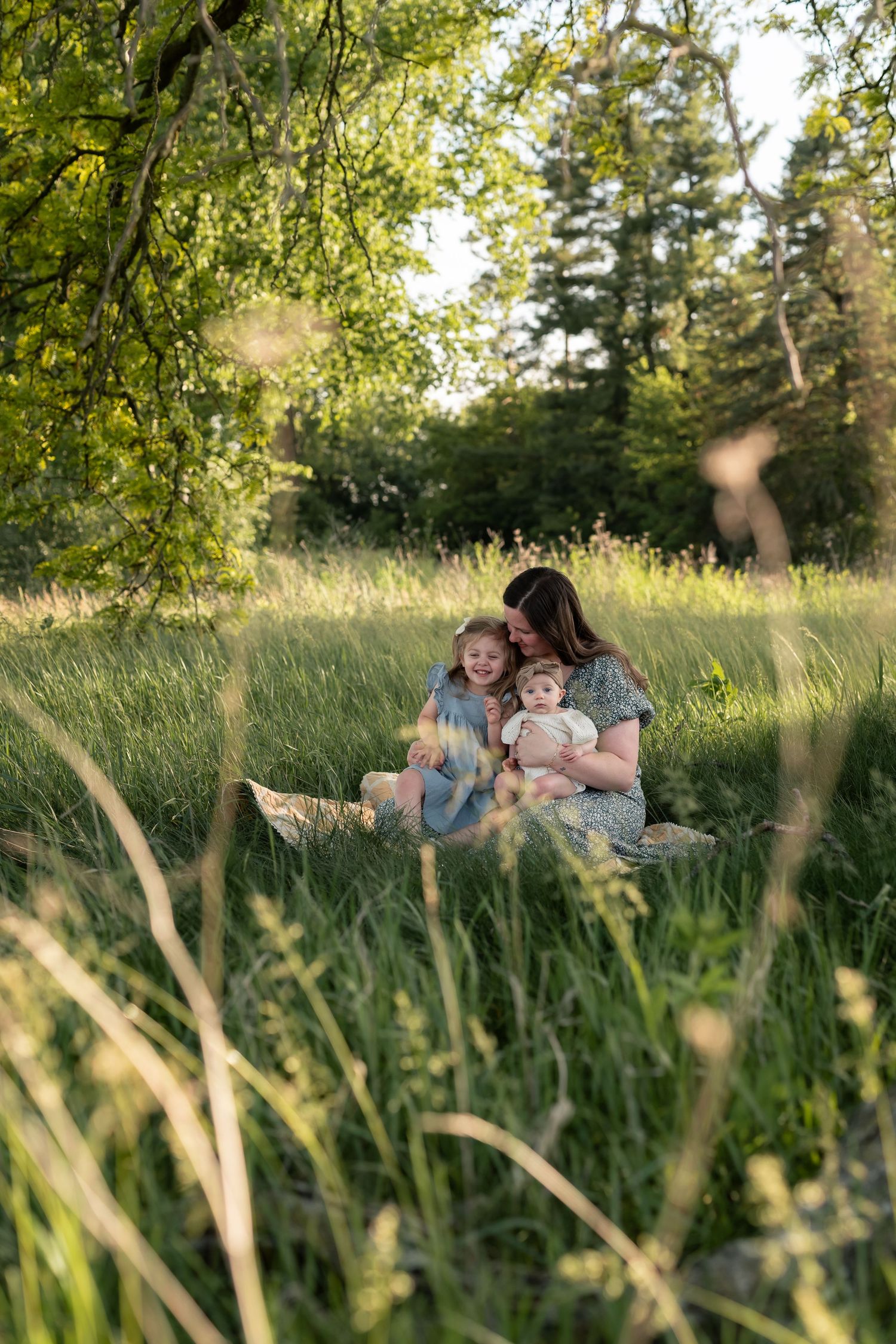 A mother sits in a lush green meadow with her two young children on a sunny day.