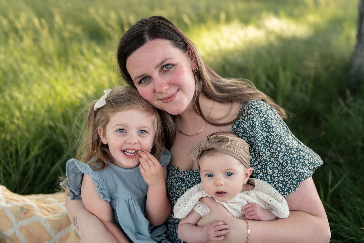 A smiling mother sits outdoors in green grass holding her baby while her young toddler daughter poses beside her.