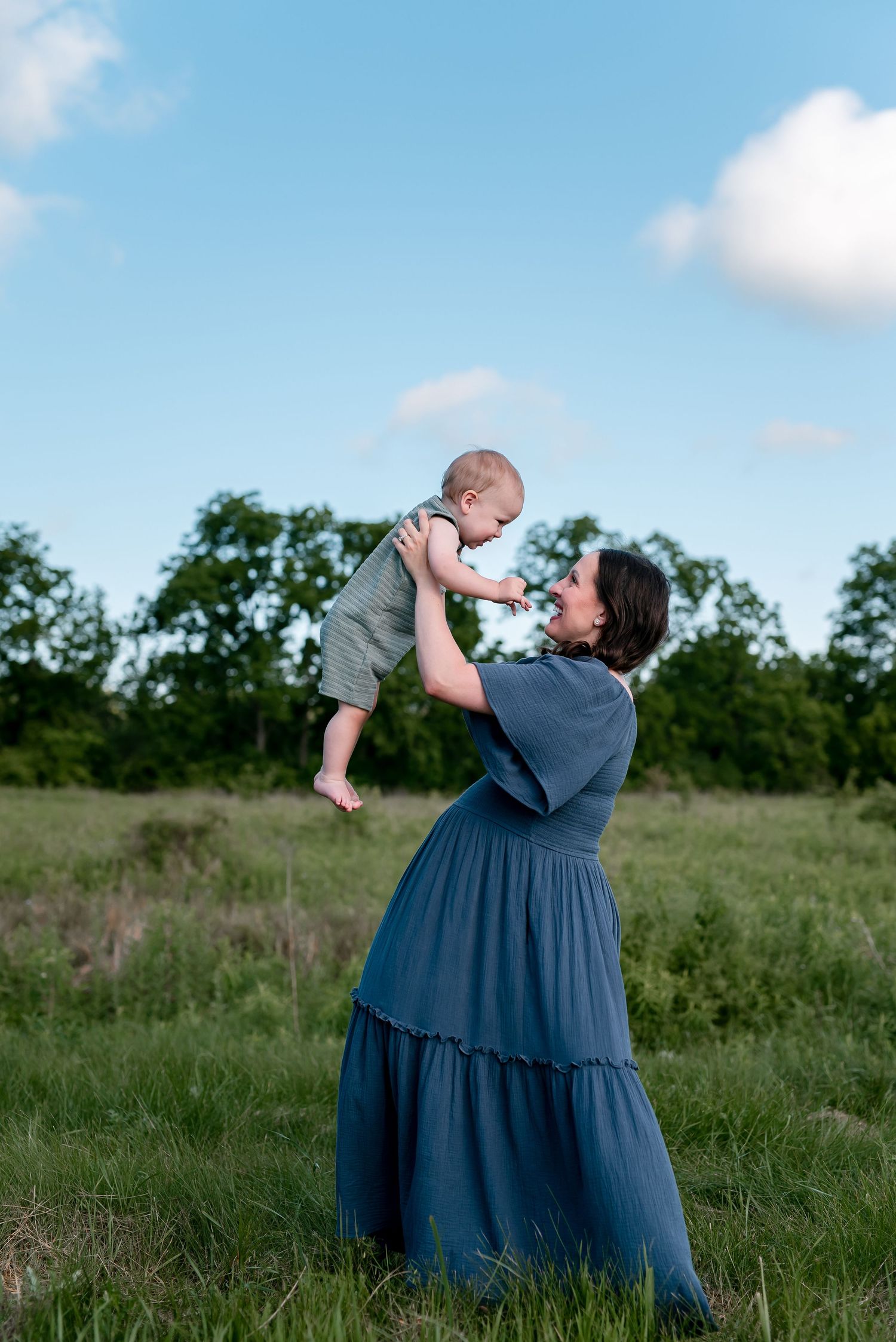 A loving moment captured in a grassy field as a blue-dressed woman lifts a baby against a bright summer sky.