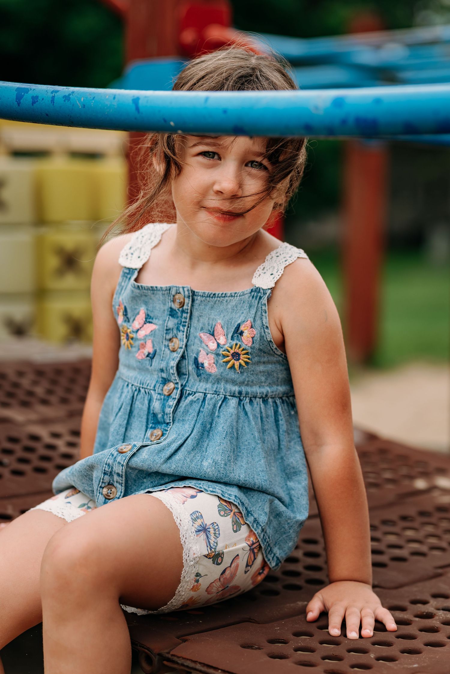 Young child in denim overalls sits on playground equipment on a warm summer day.