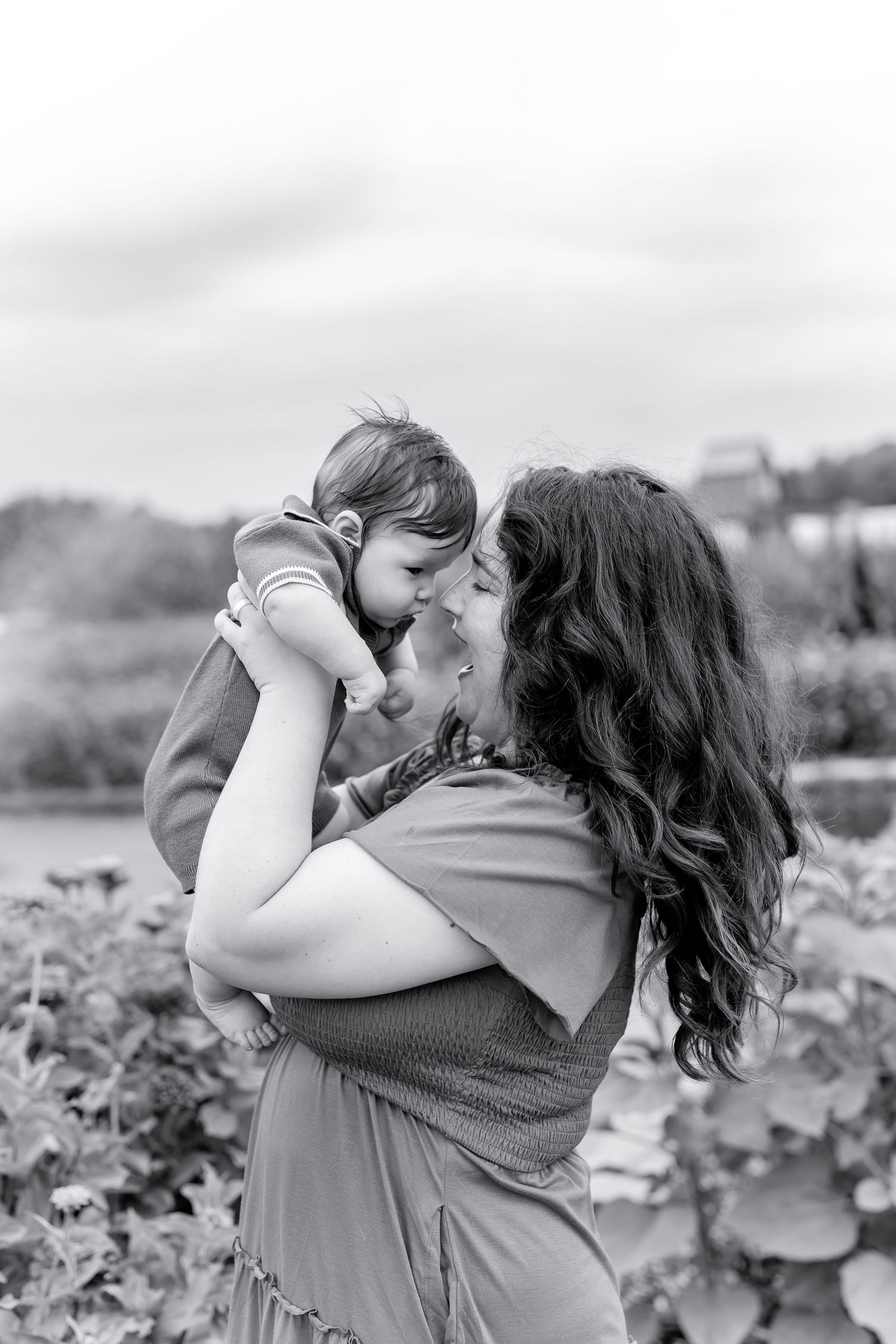 Black and white photo of a mother lifting and kissing her baby outdoors among green plants.