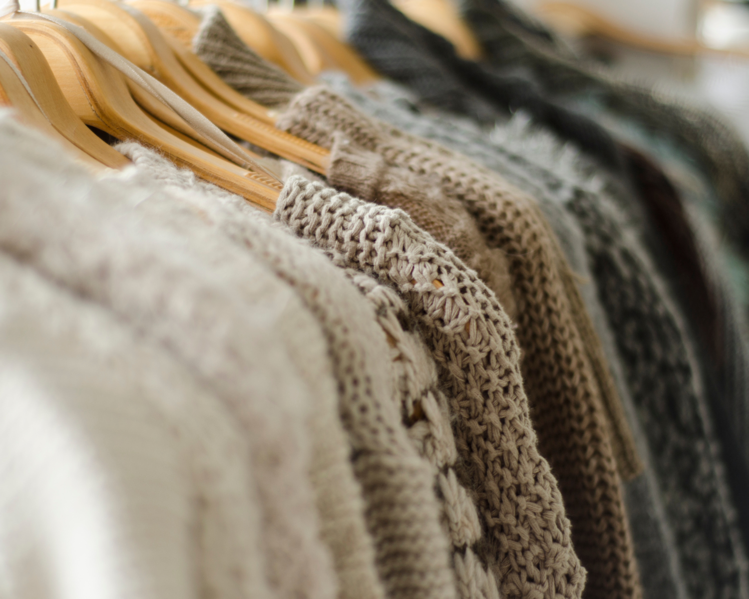 A row of cozy knit sweaters in neutral tones hanging on wooden hangers on a clothing rack.