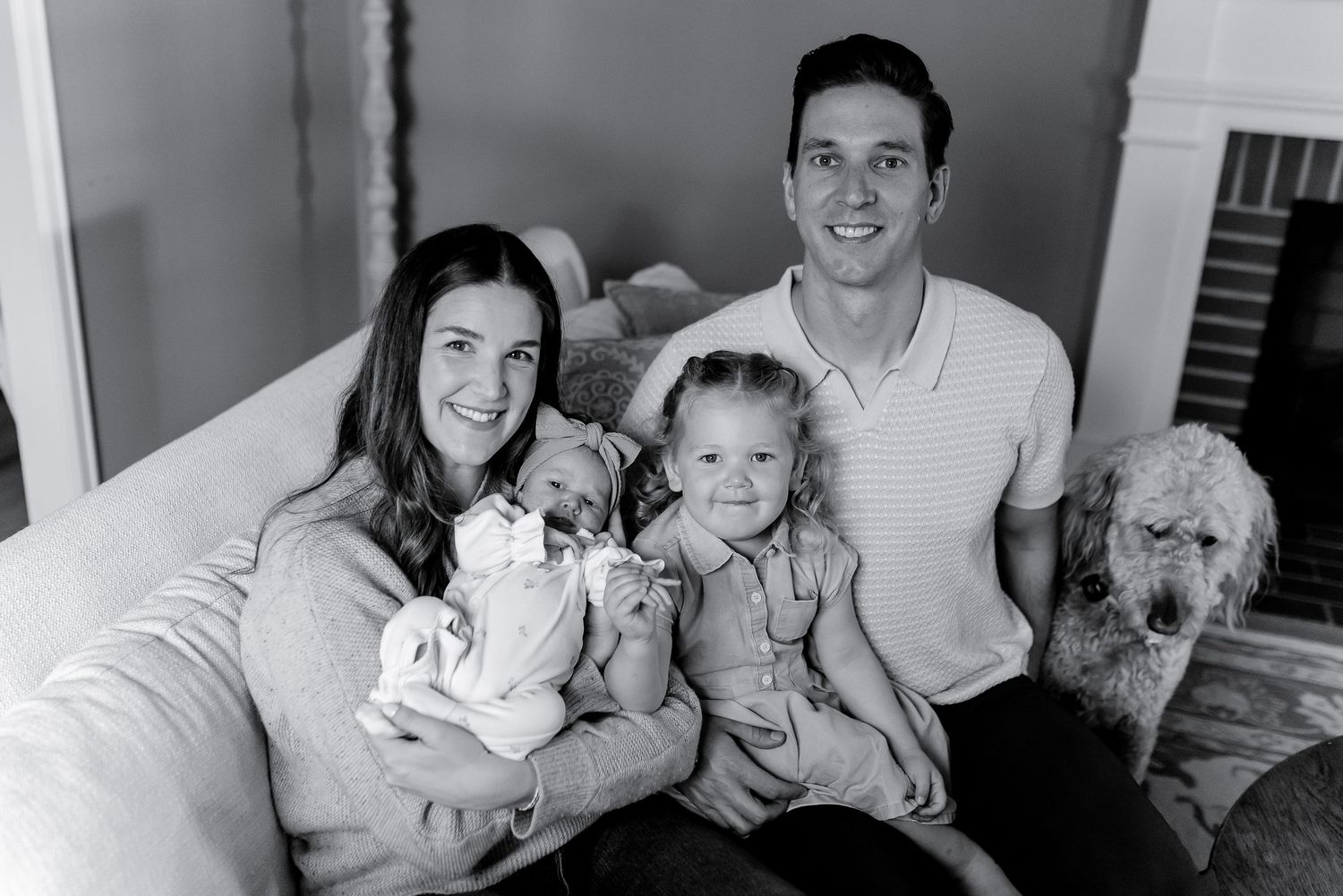 Black and white family portrait with parents, newborn, toddler girl, and fluffy dog on couch.