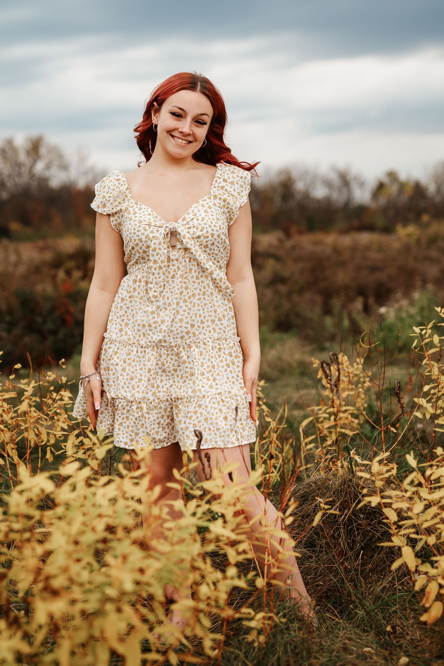Smiling redhead woman in floral ruffle dress posing in golden autumn meadow.