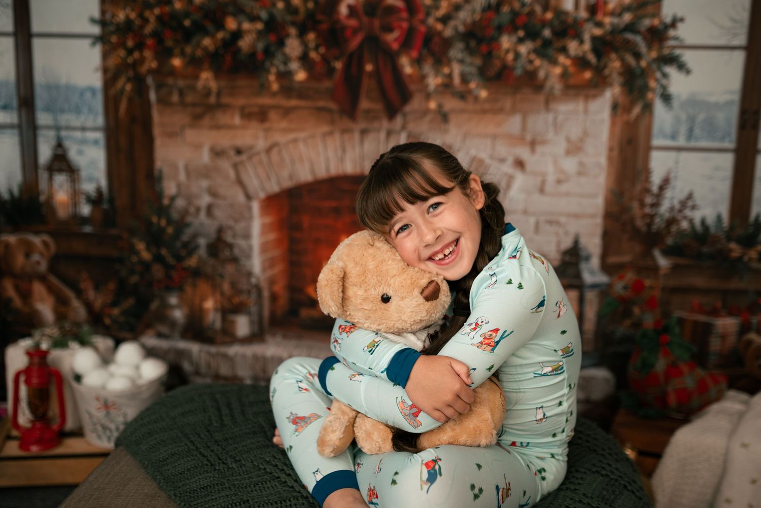Smiling girl in holiday pajamas hugging a teddy bear in front of a decorated brick fireplace with garland and Christmas ornaments.