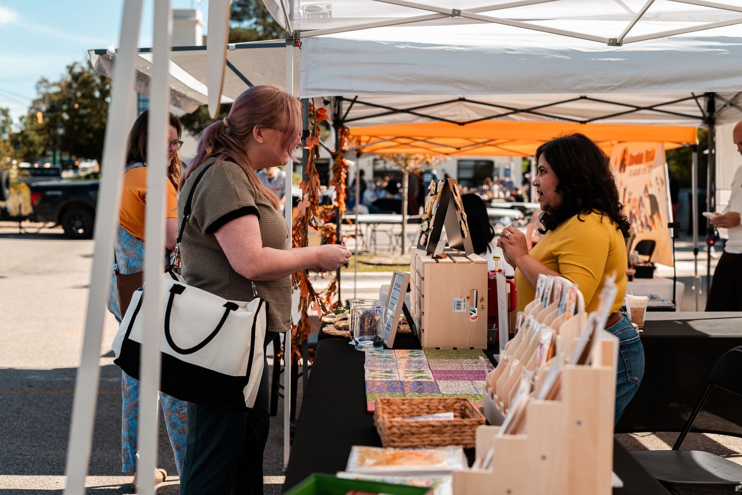 Two women browsing handmade goods at a sunny outdoor artisan market booth under a white canopy.