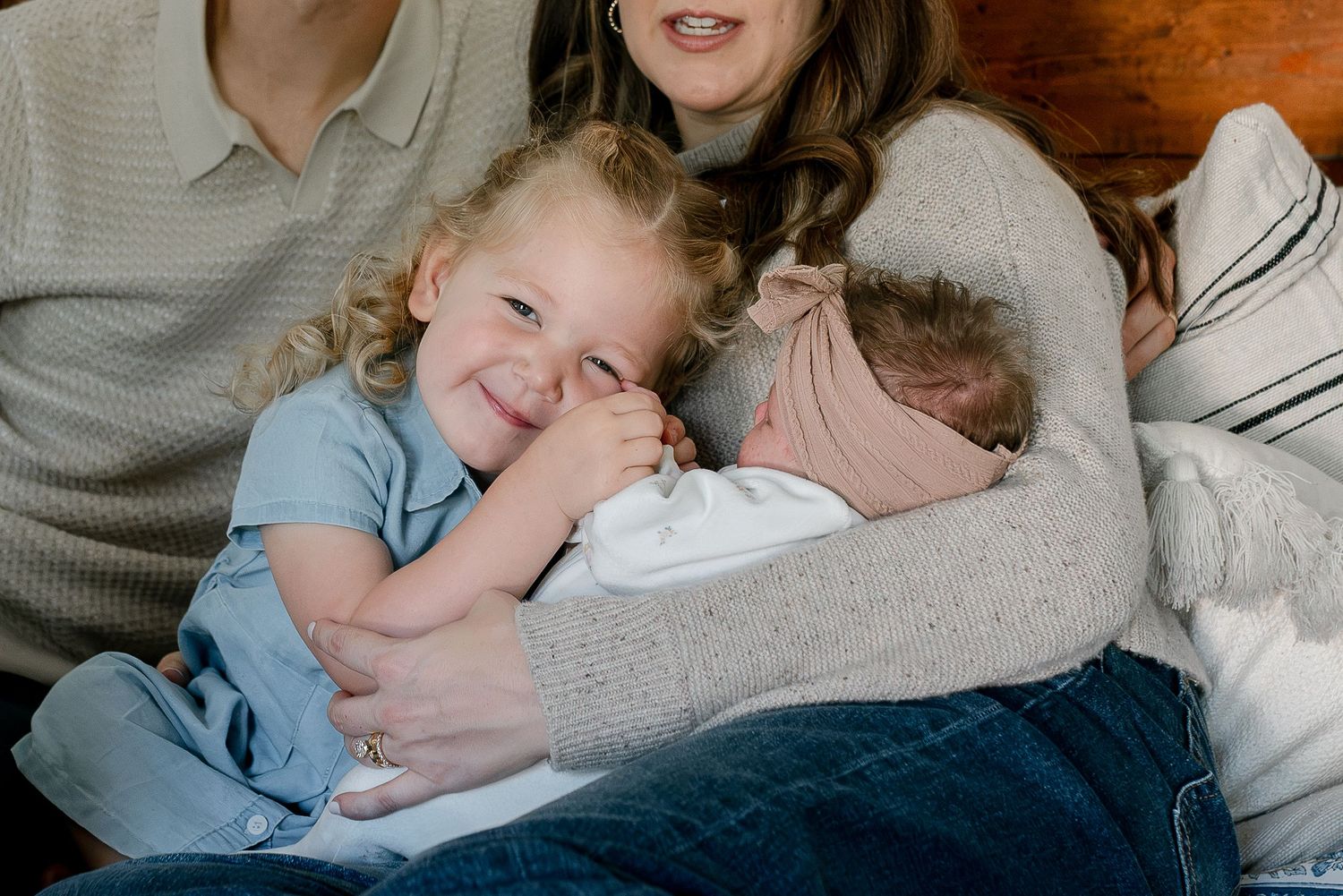 A toddler smiles while holding a newborn baby, as a mother cradles both children in a warm family portrait.