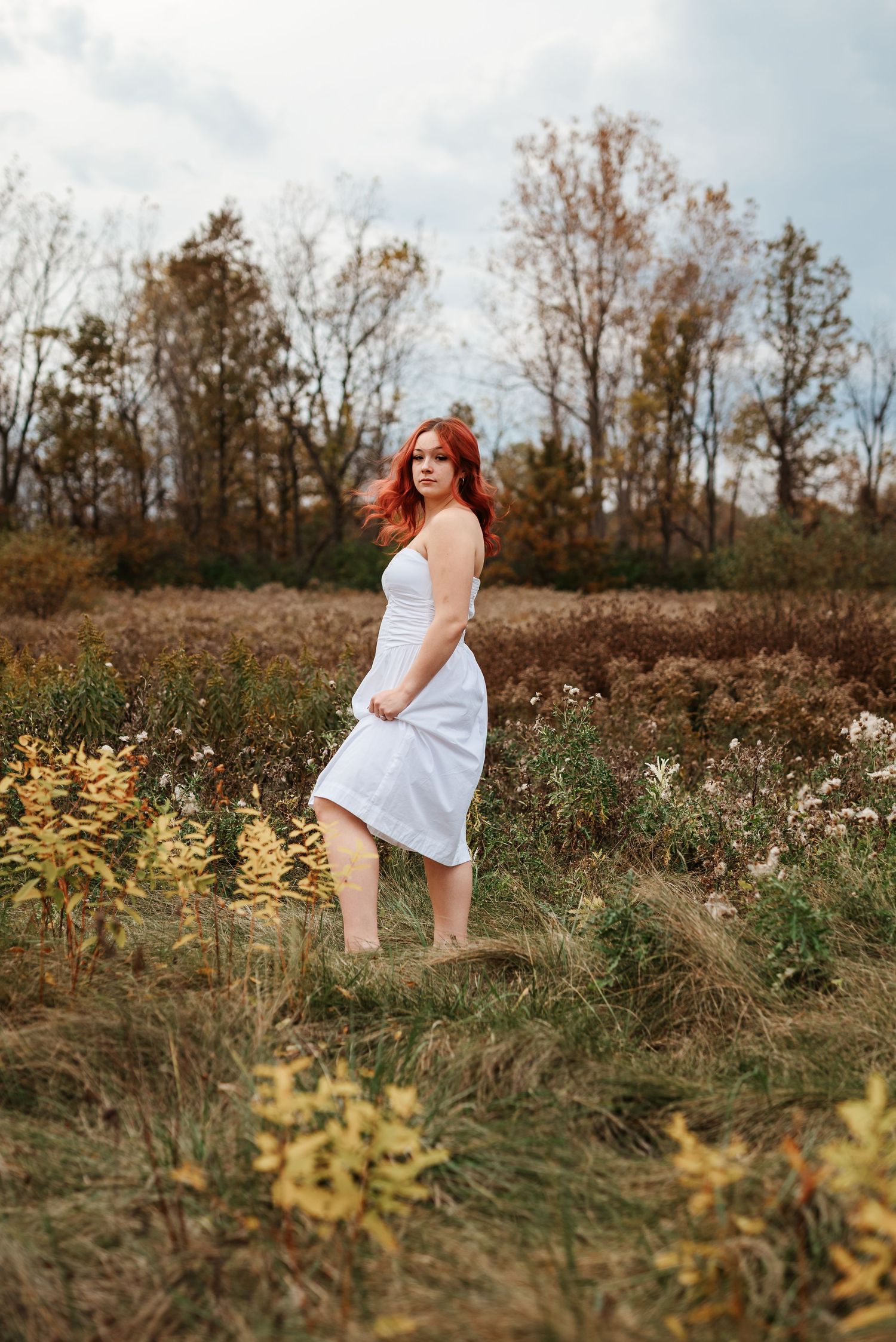 Red-haired woman in white midi dress standing in autumn field with wildflowers and bare trees.