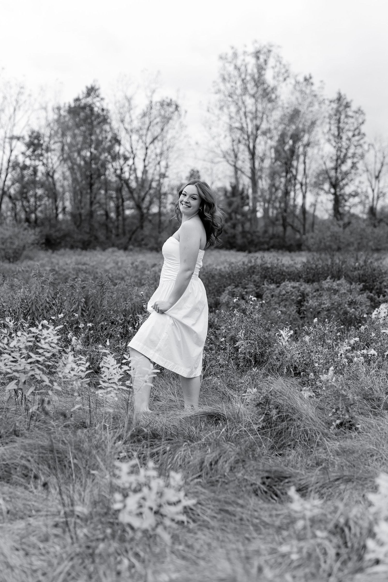 Black and white photo of smiling young woman in white dress twirling in a wildflower field.