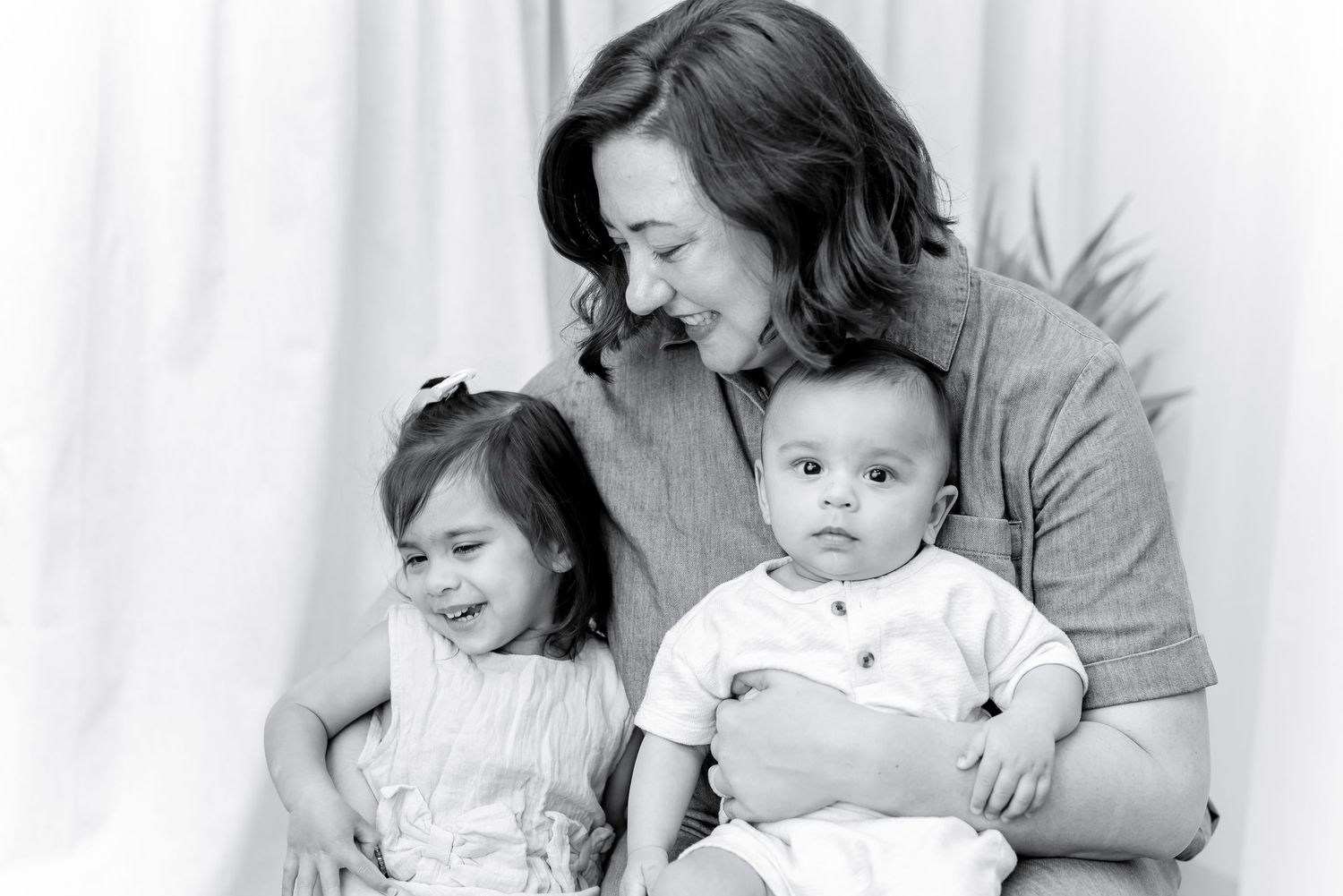 Black and white portrait of a mother smiling while holding her baby and young daughter in a bright studio setting.