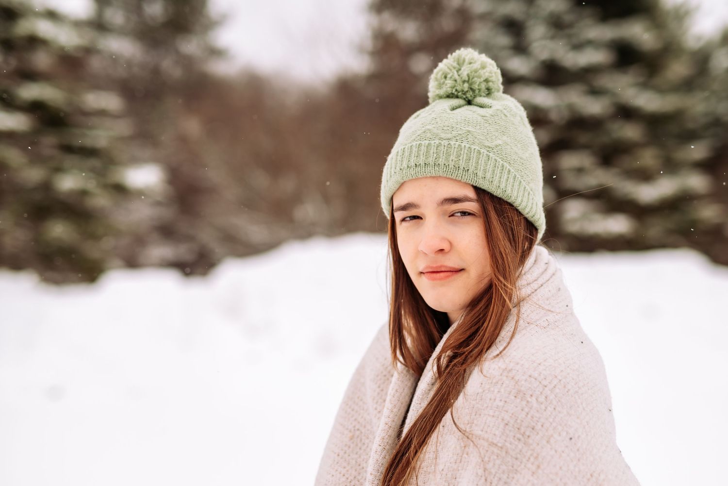 A person wearing a knit beanie and winter coat poses in a snowy winter landscape with evergreen trees in the background.