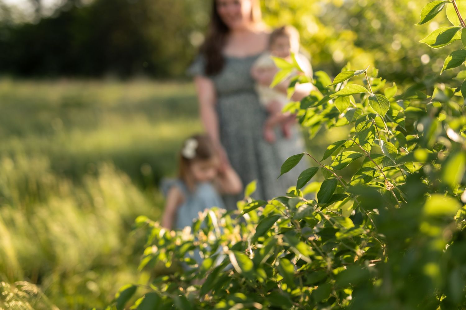 Mother holding baby with young daughter in sunlit green garden, shallow depth of field with lush foliage foreground.