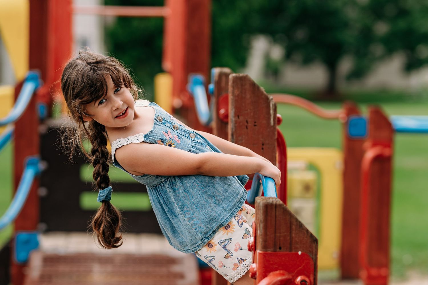 Young child playing on red and blue playground equipment in a park during a sunny day.