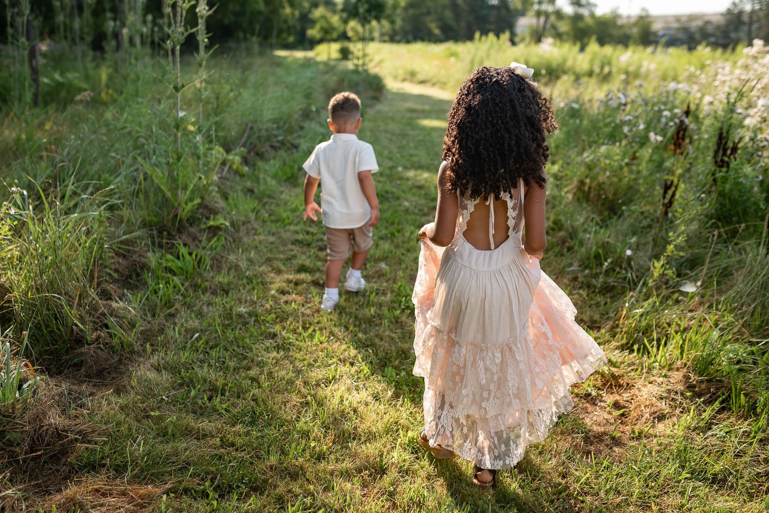Two children walking through a lush green meadow path, girl in pink dress with curly hair, boy in white shirt.