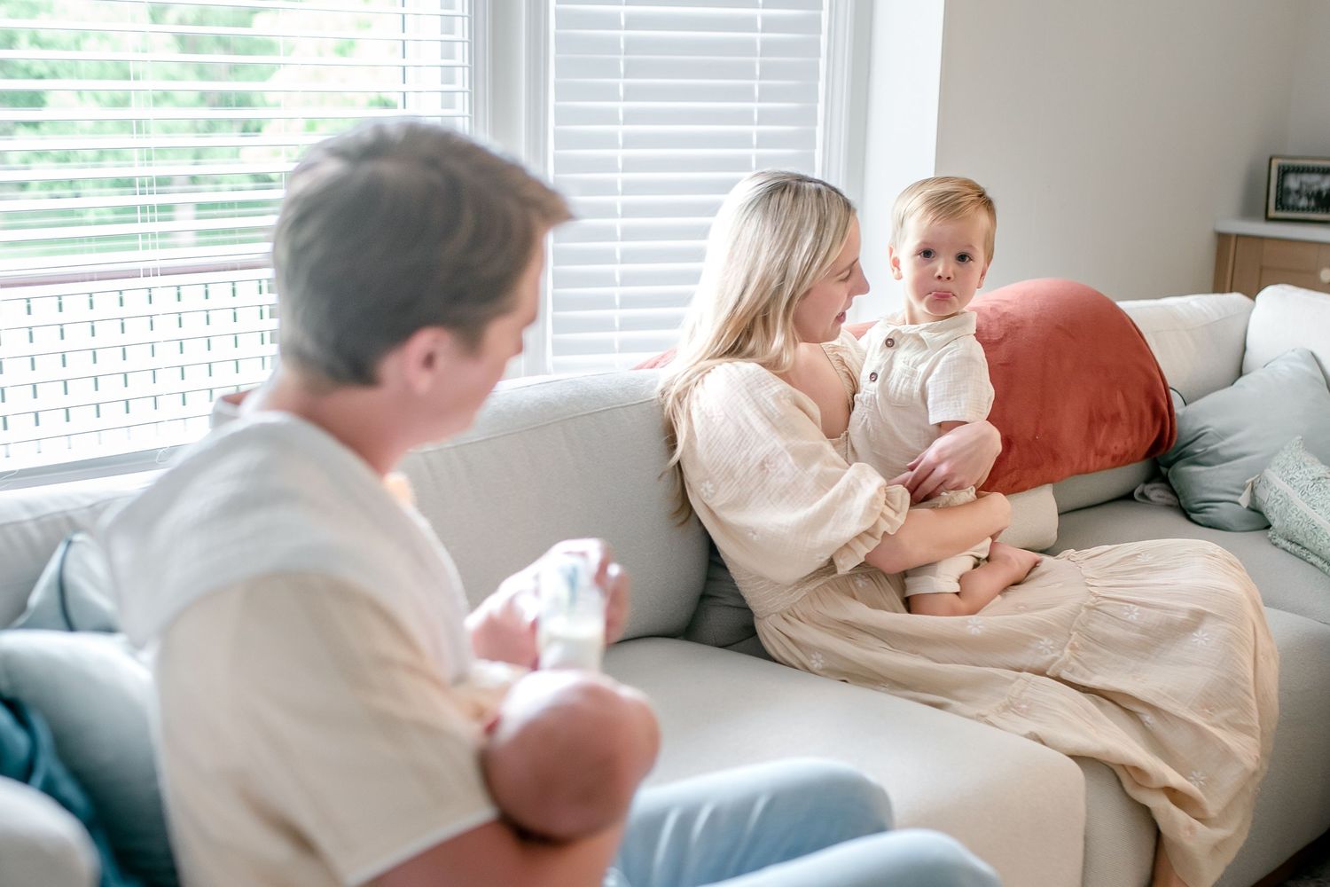 Family sitting on couch with mother holding toddler and father cradling newborn baby in bright living room.