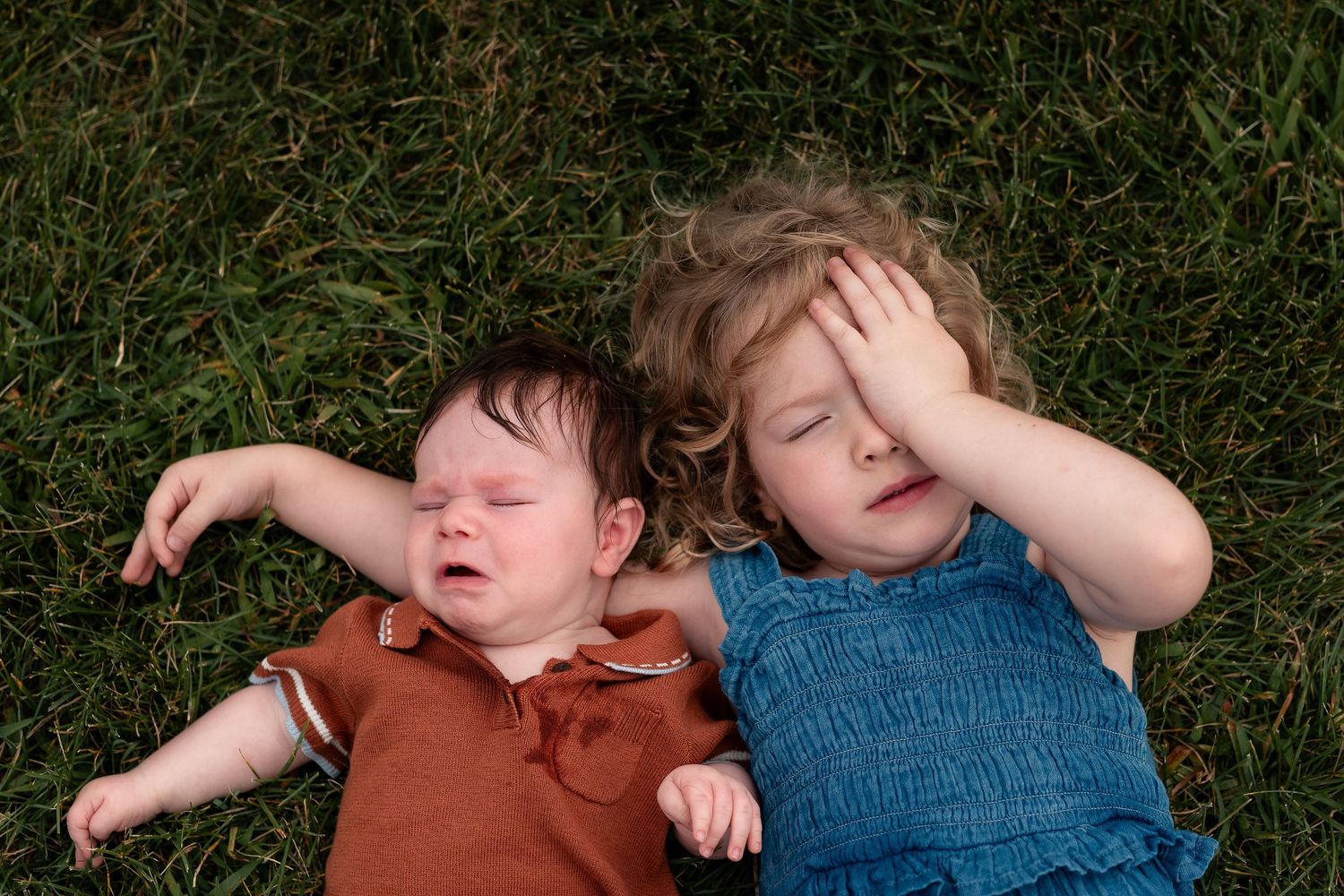 Two young siblings lying on green grass, baby in rust romper and toddler girl in blue dress resting.
