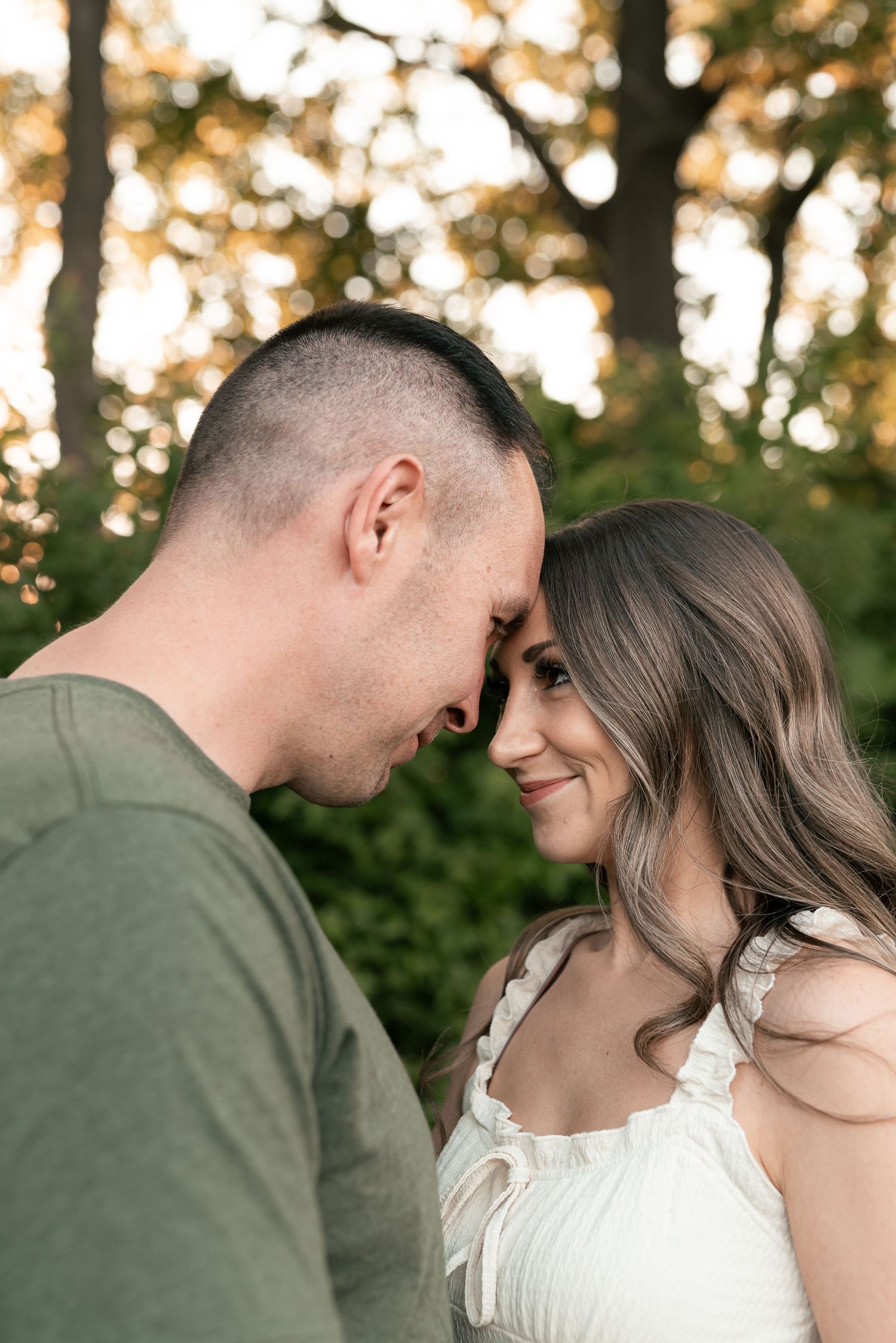Couple sharing an intimate forehead touch outdoors, woman in white dress smiling softly, man in green shirt, golden bokeh trees.