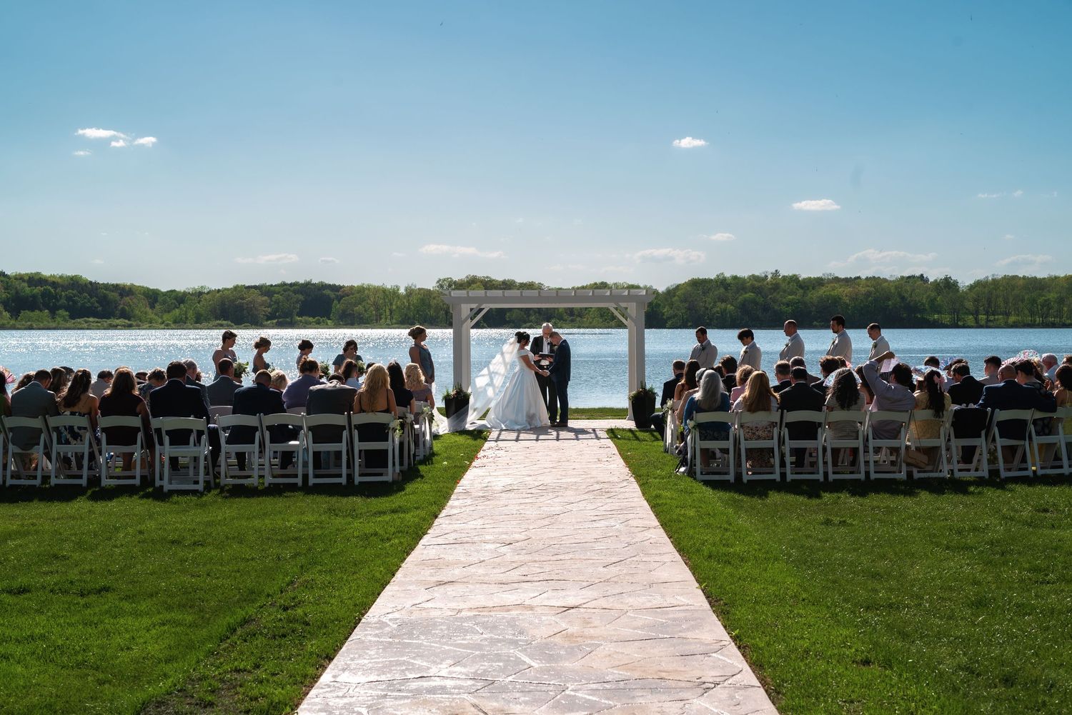 A lakeside outdoor wedding ceremony setup with white chairs and aisle runner on green grass overlooking the water.