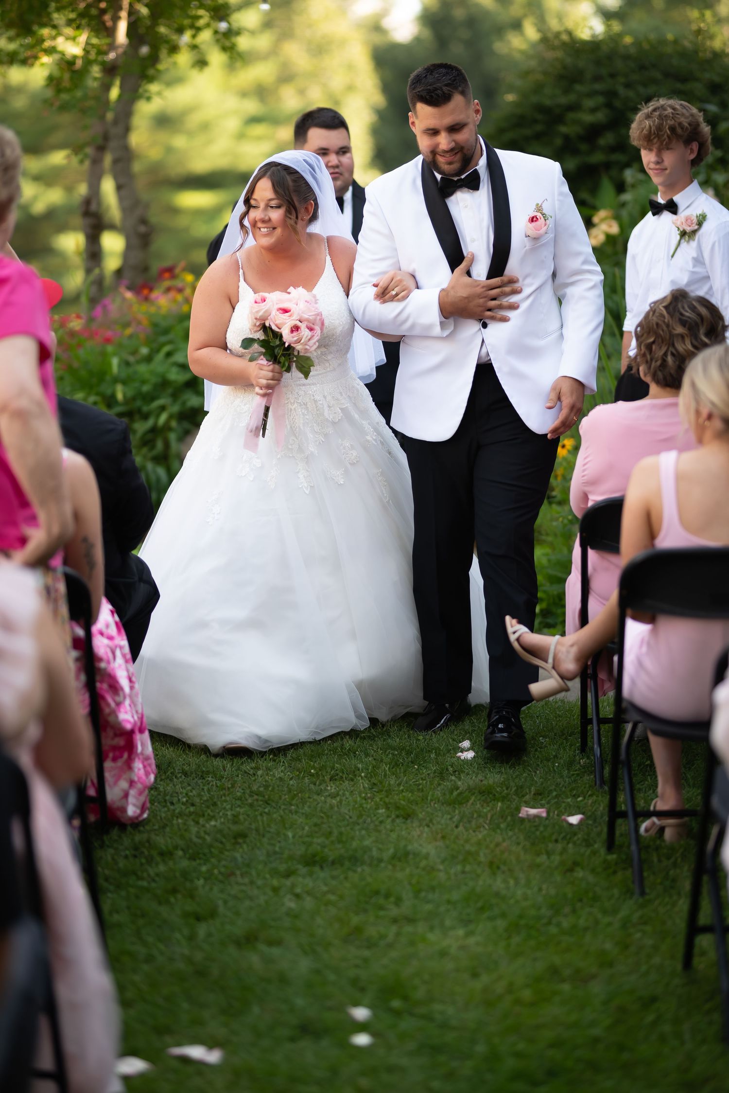 Newlywed couple walks down outdoor garden aisle after ceremony with guests in pink and black attire looking on.