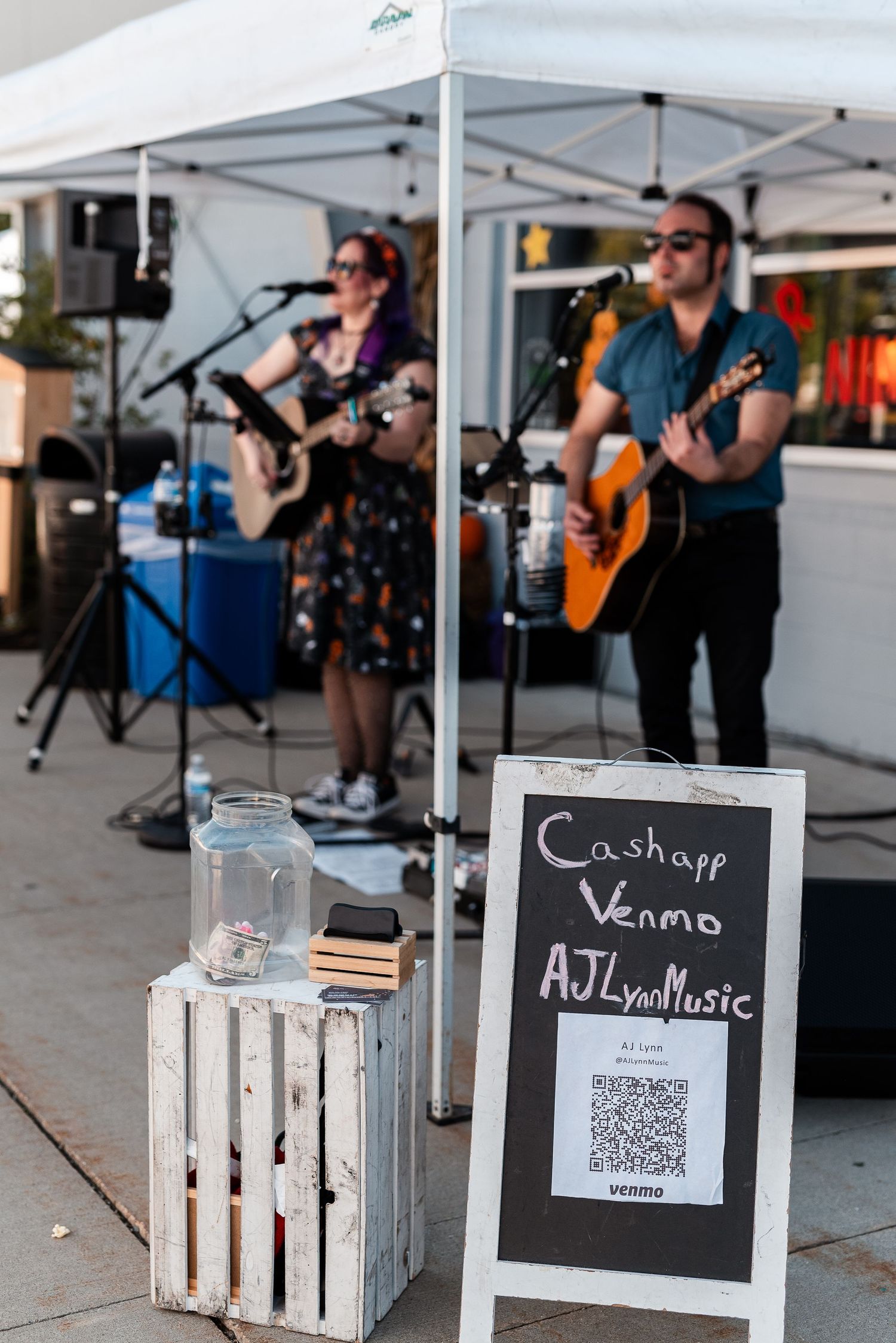 AJLyn Music duo performs live acoustic set outdoors with Venmo QR code sign on white wooden crate.