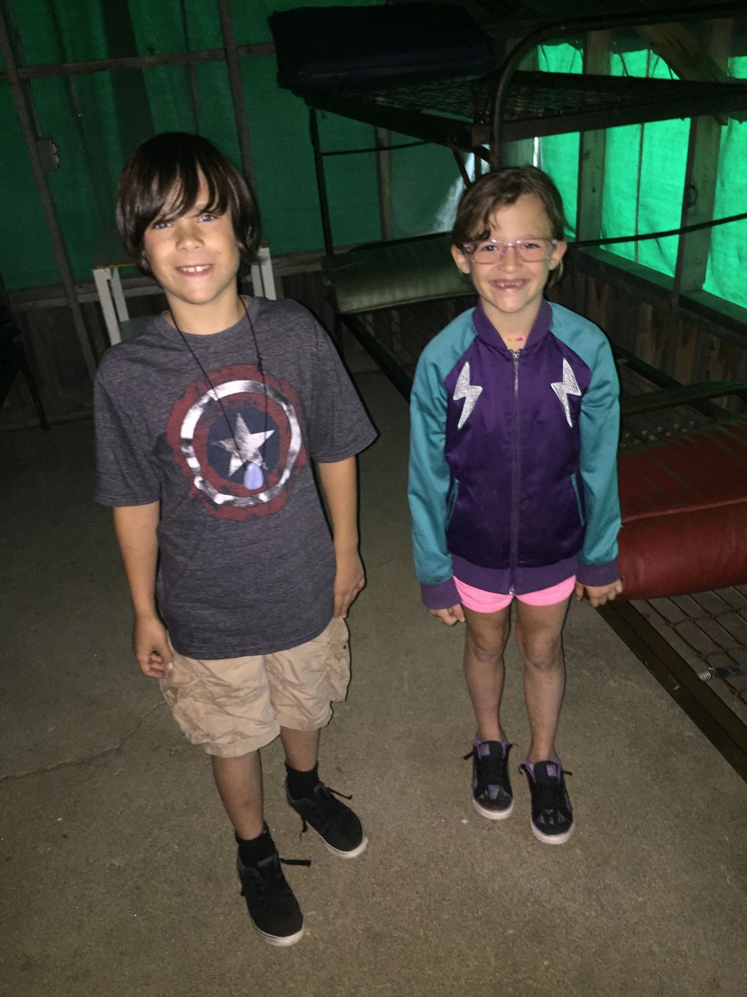 Two smiling children pose indoors, boy in Captain America shirt and khaki shorts, girl in purple jacket and pink shorts.
