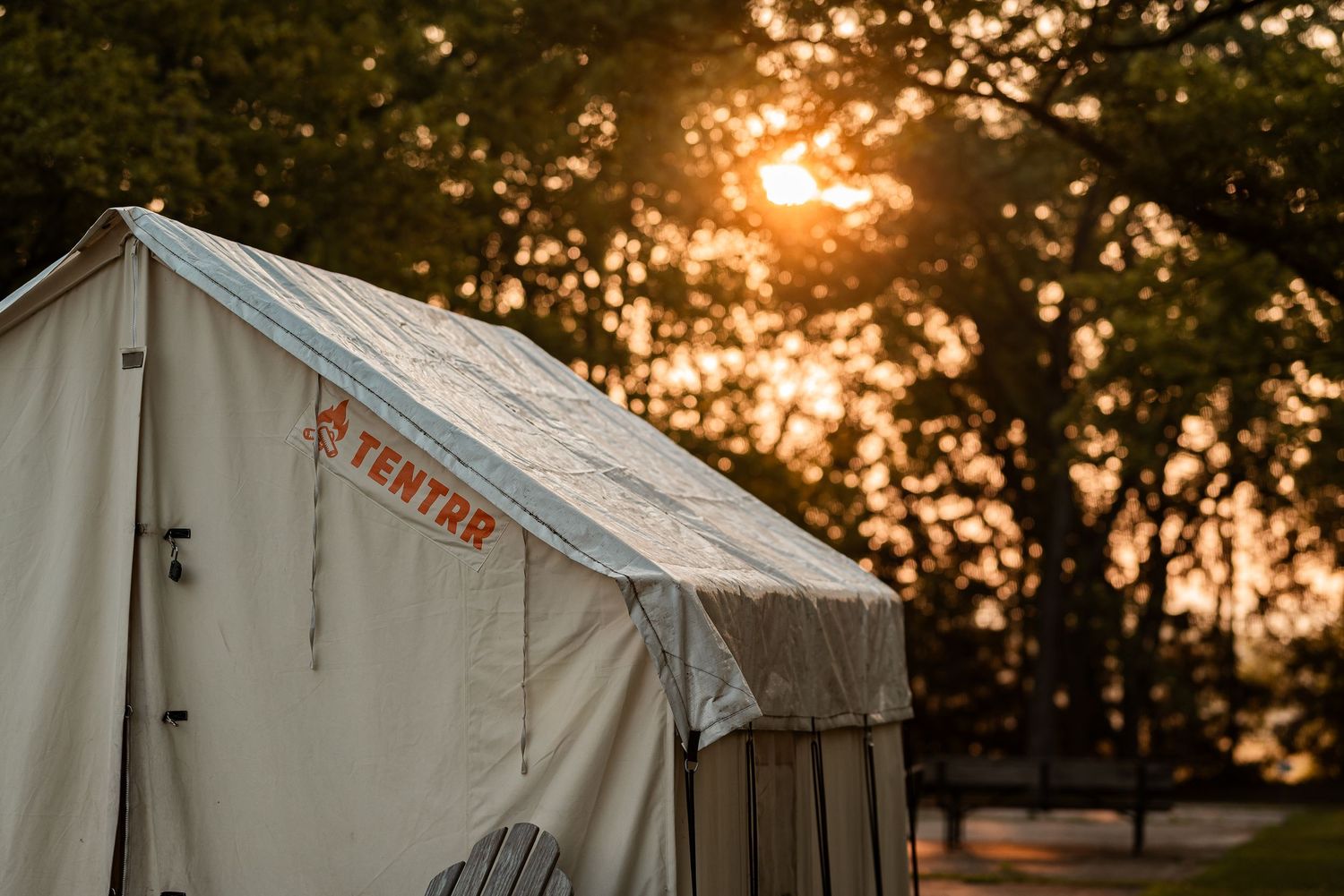 A Tentrr canvas tent glows warmly at sunset, surrounded by tall trees in a scenic campsite setting.