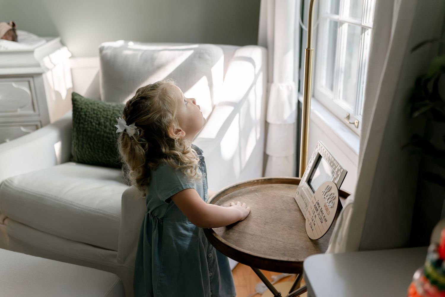 Young girl in blue dress looking up at framed photo on round wooden table by sunny window.