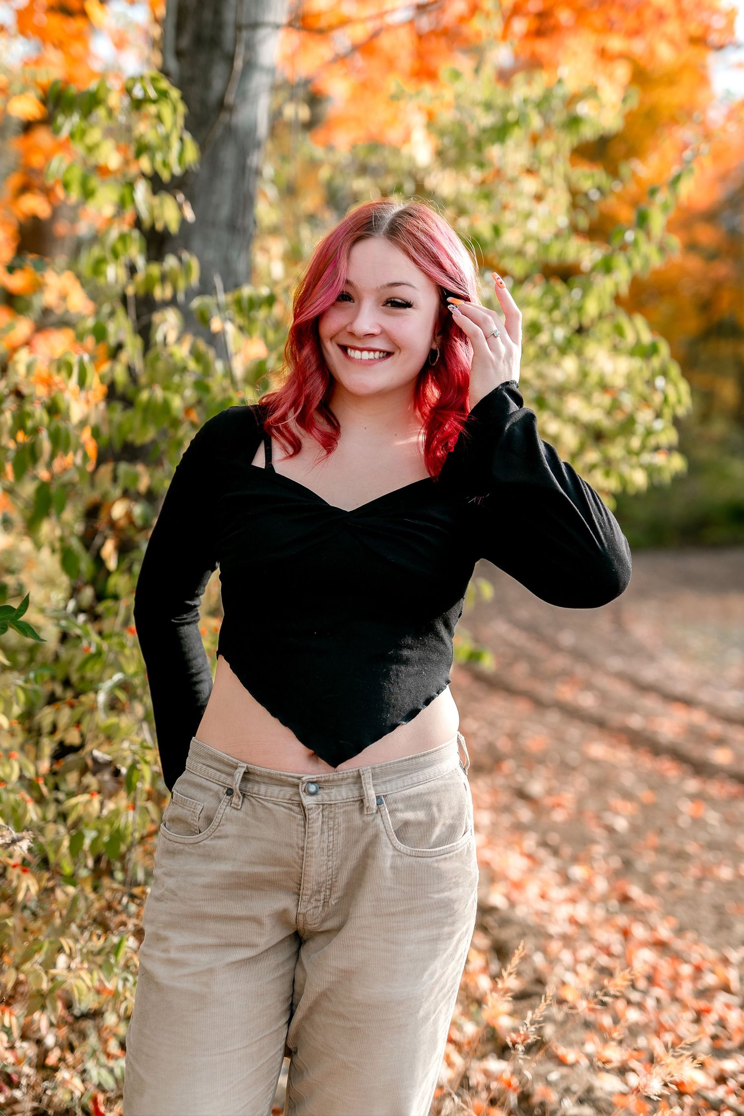 Young woman with red hair in black top and khaki jeans smiling outdoors among colorful autumn foliage.