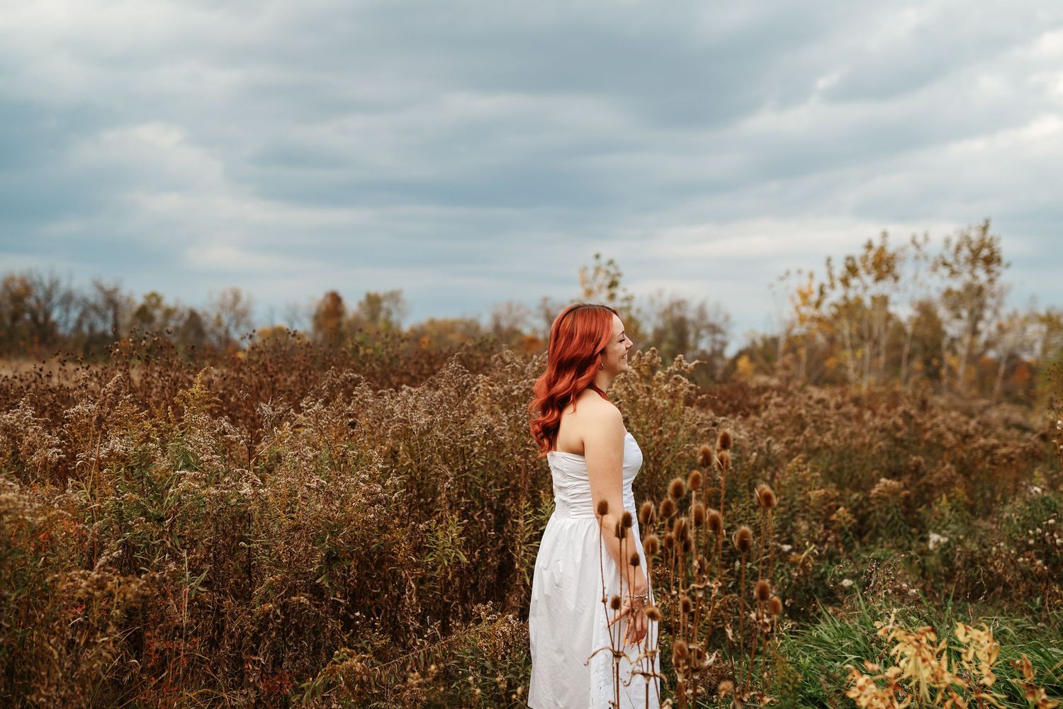 A red-haired woman in a white dress stands in an autumn field under a cloudy sky.