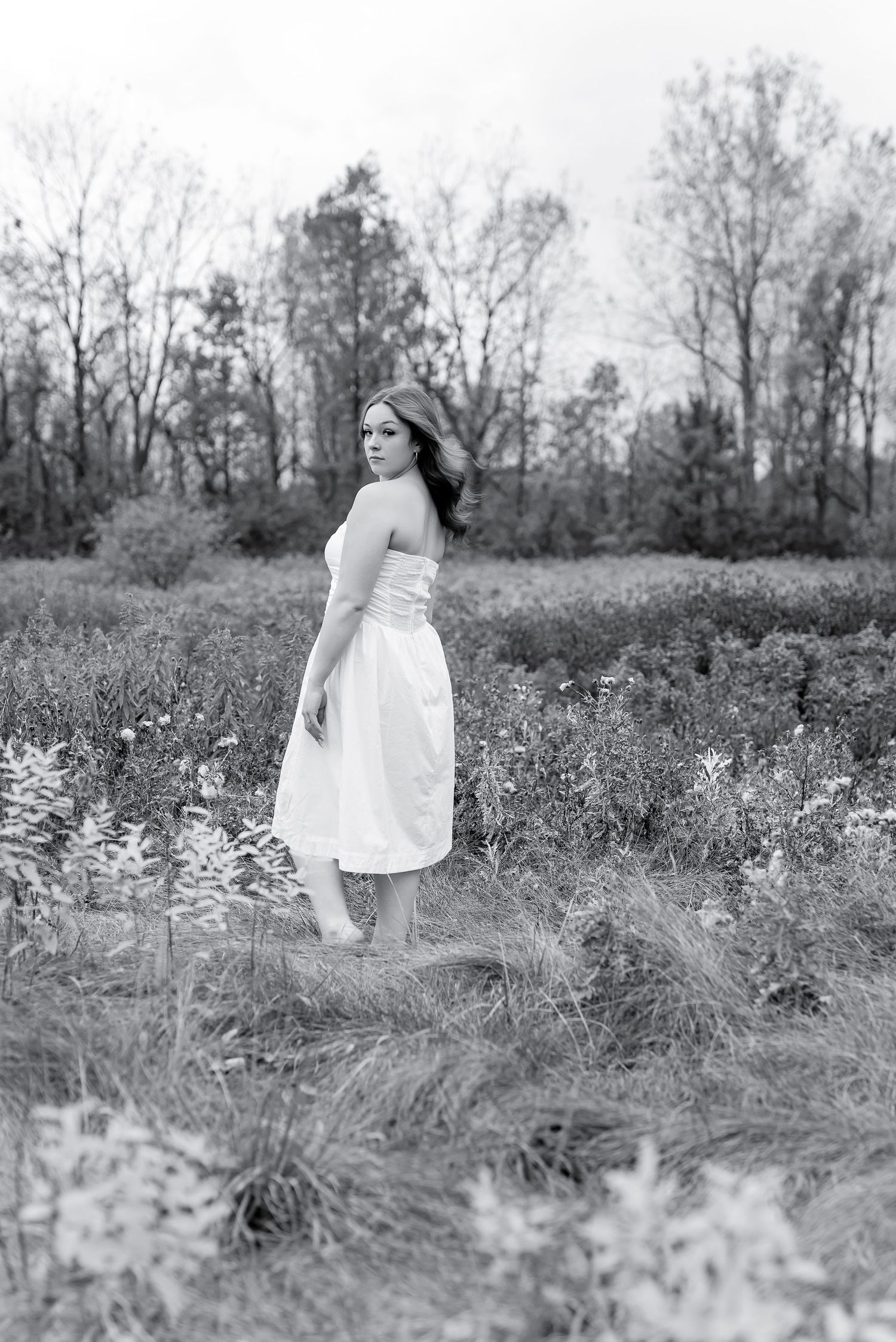 Woman in white midi dress standing in overgrown field surrounded by wildflowers and trees.