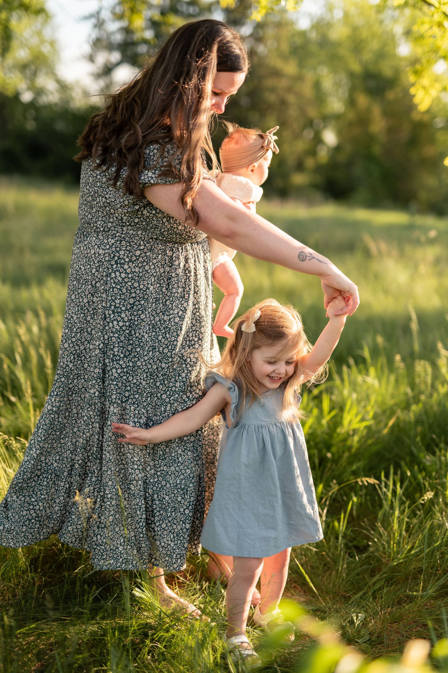 A mother in a floral dress holds her baby while dancing with her toddler daughter in a sunny green meadow.