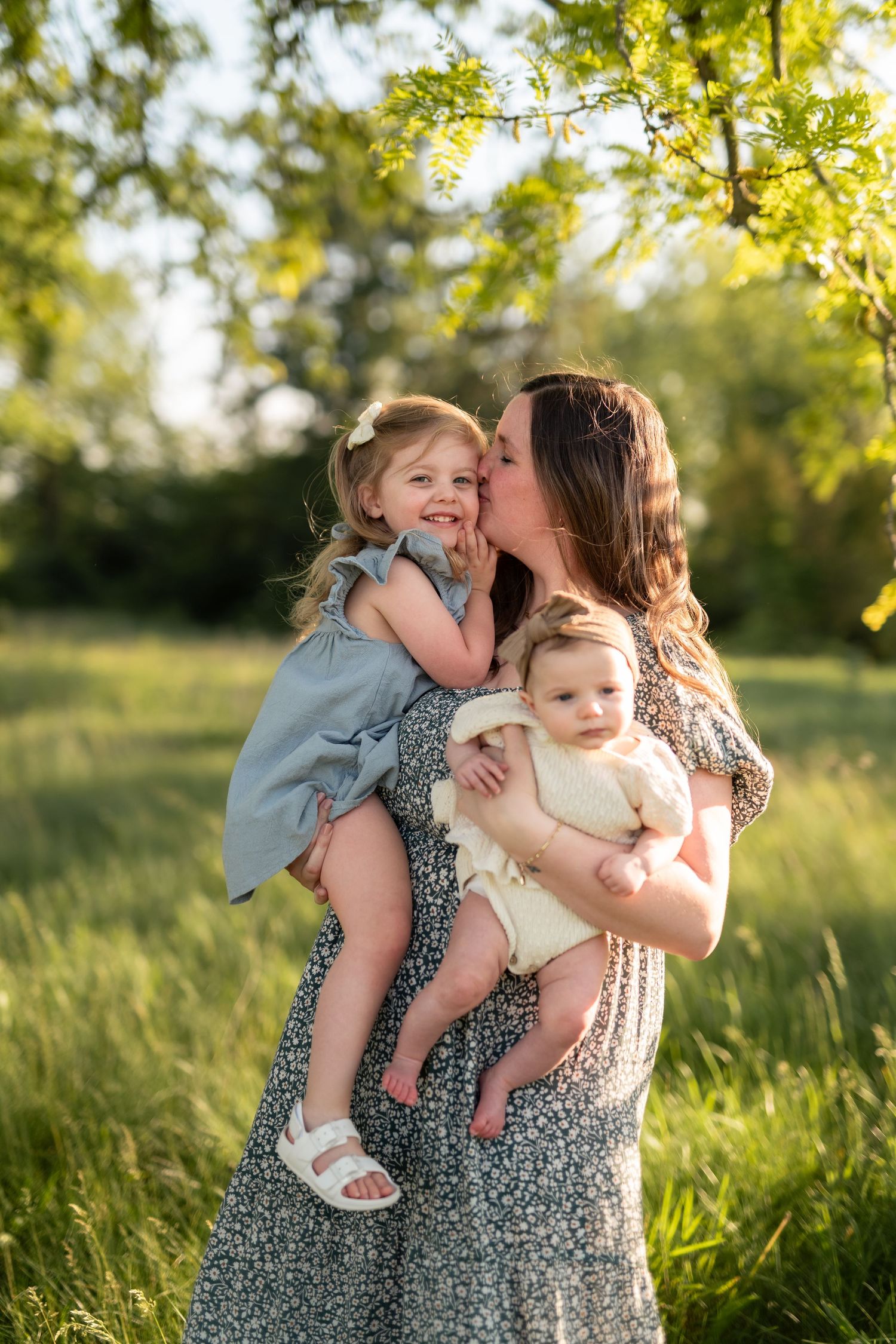 A mother kisses her toddler daughter while holding a baby in a sunny green field.