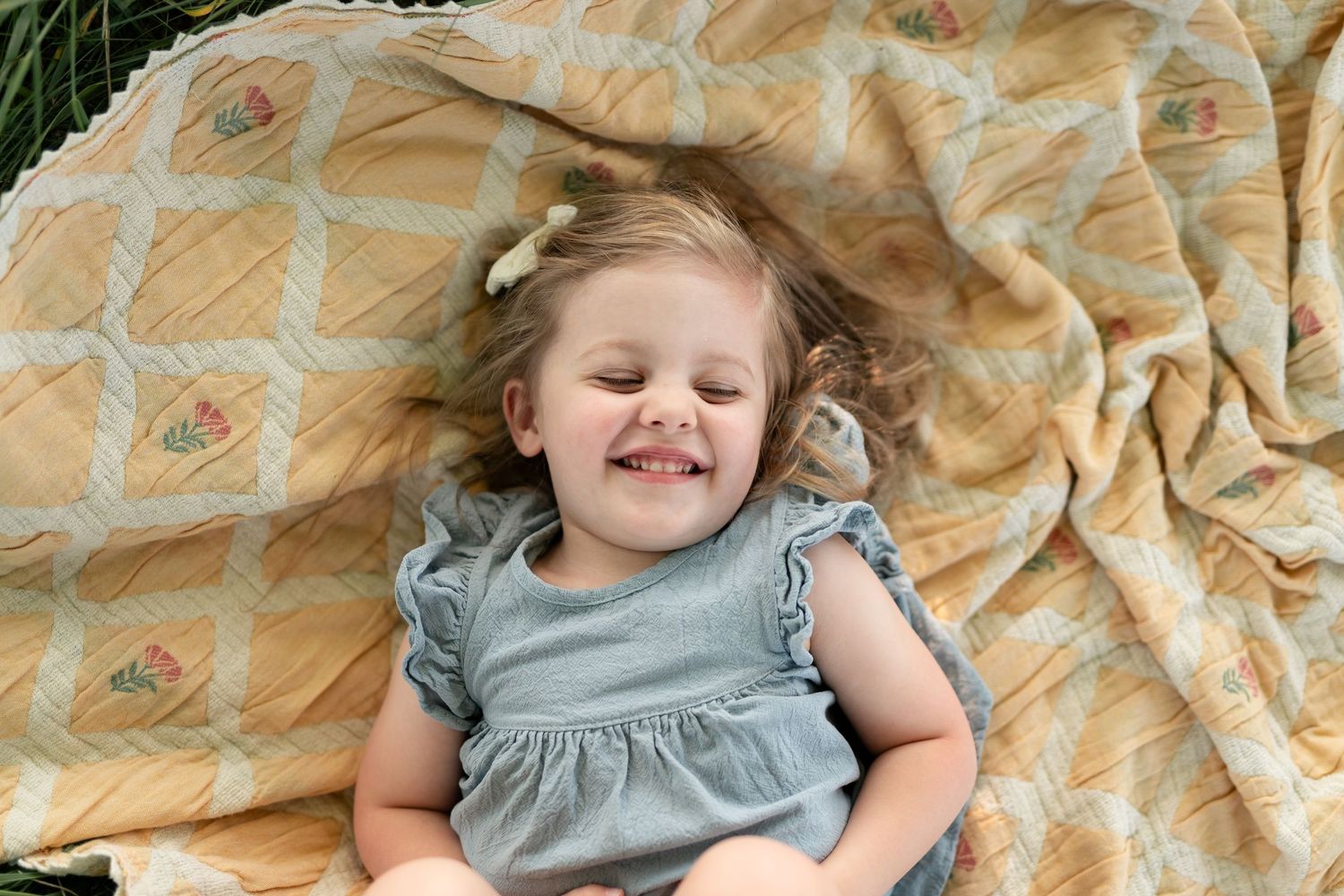 Smiling toddler girl with hair bow lying on a yellow patterned blanket outdoors in a gray ruffled dress.