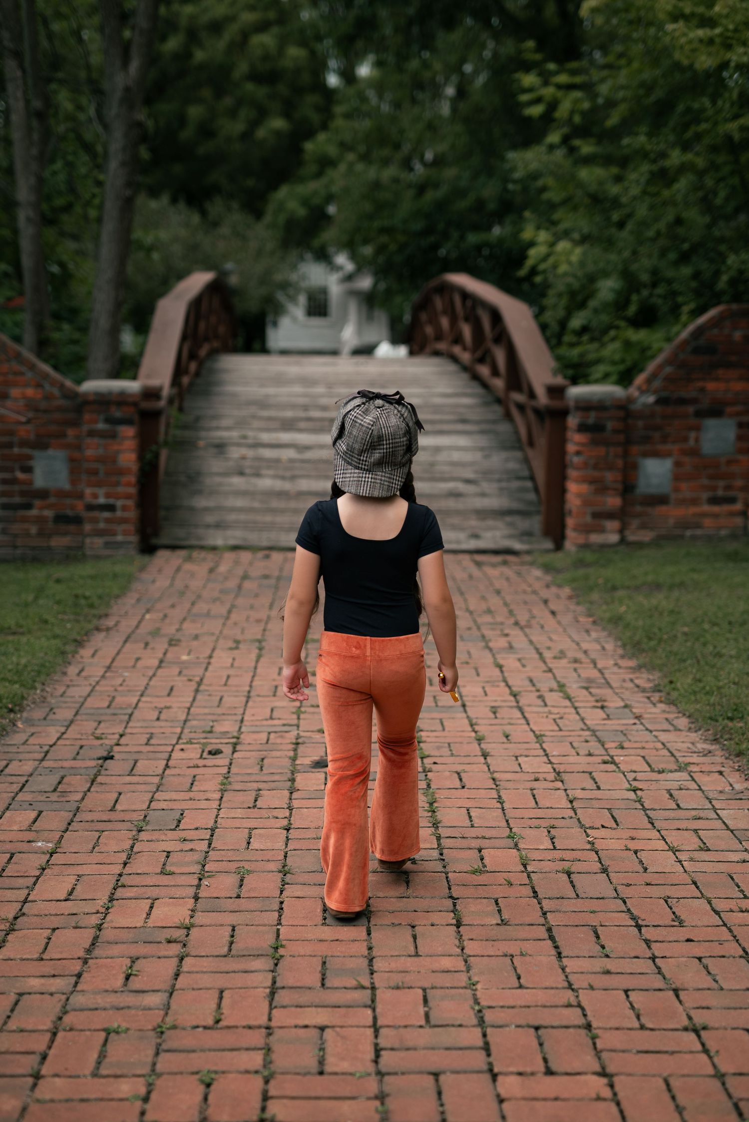 Child in orange flare pants and plaid hat walks along brick path toward arched bridge surrounded by green trees.