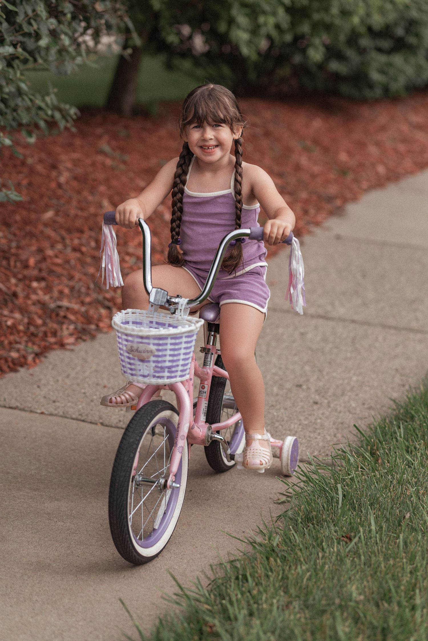 A child in a purple tank top rides a pink bicycle with streamers and a white basket on a sidewalk near mulch.