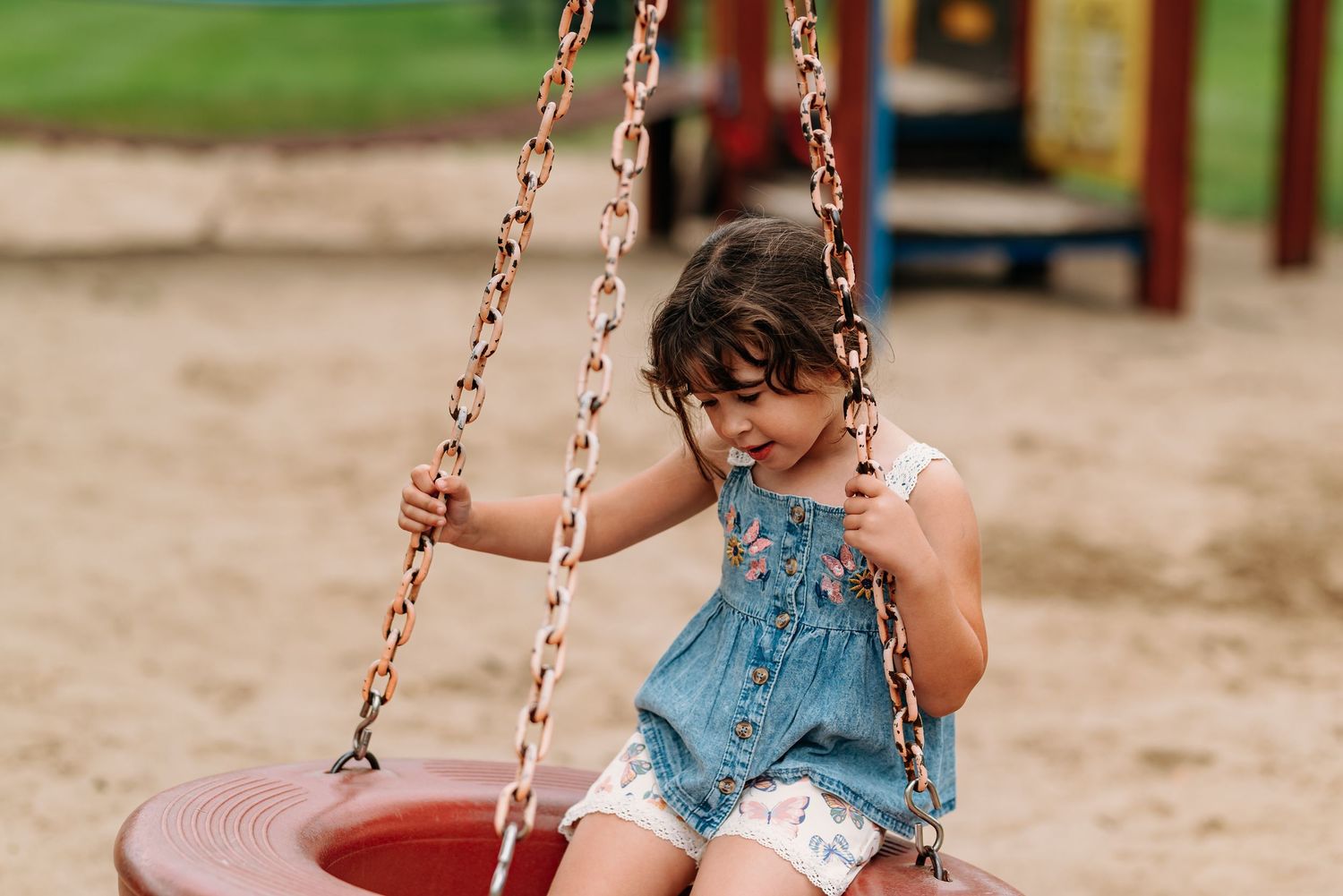Young child in a blue dress sitting on a red disc swing at a playground during summer.