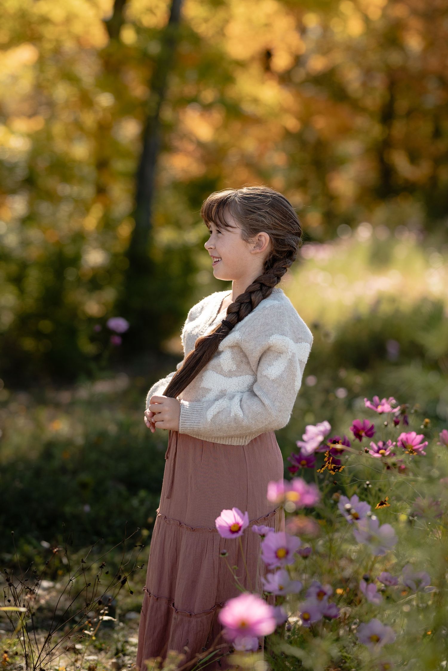 Smiling girl with a long braid in a cream cardigan and rose skirt stands among pink wildflowers in a golden autumn meadow.