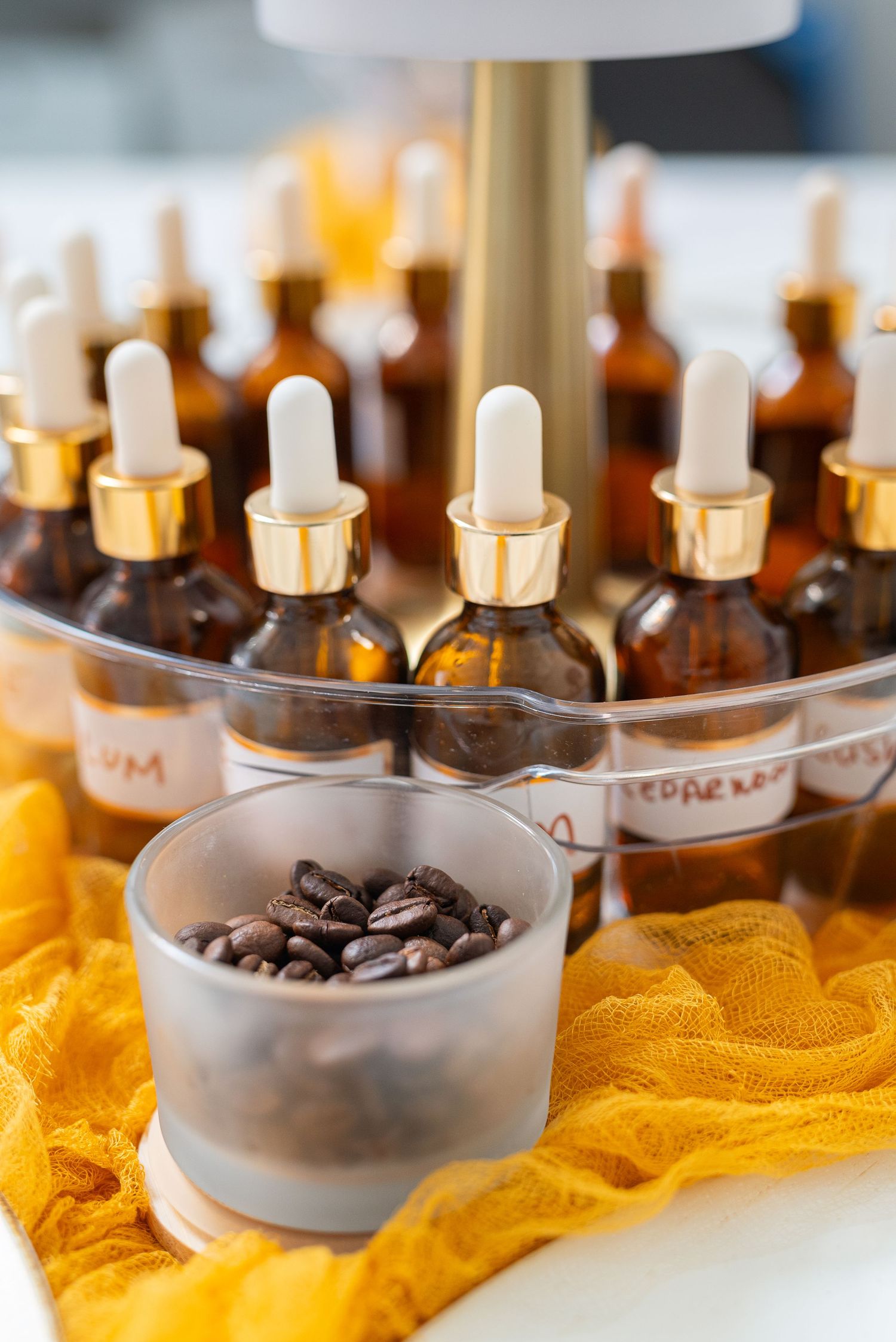 Amber dropper bottles with gold caps displayed on a tiered stand with coffee beans in a glass bowl on yellow fabric.