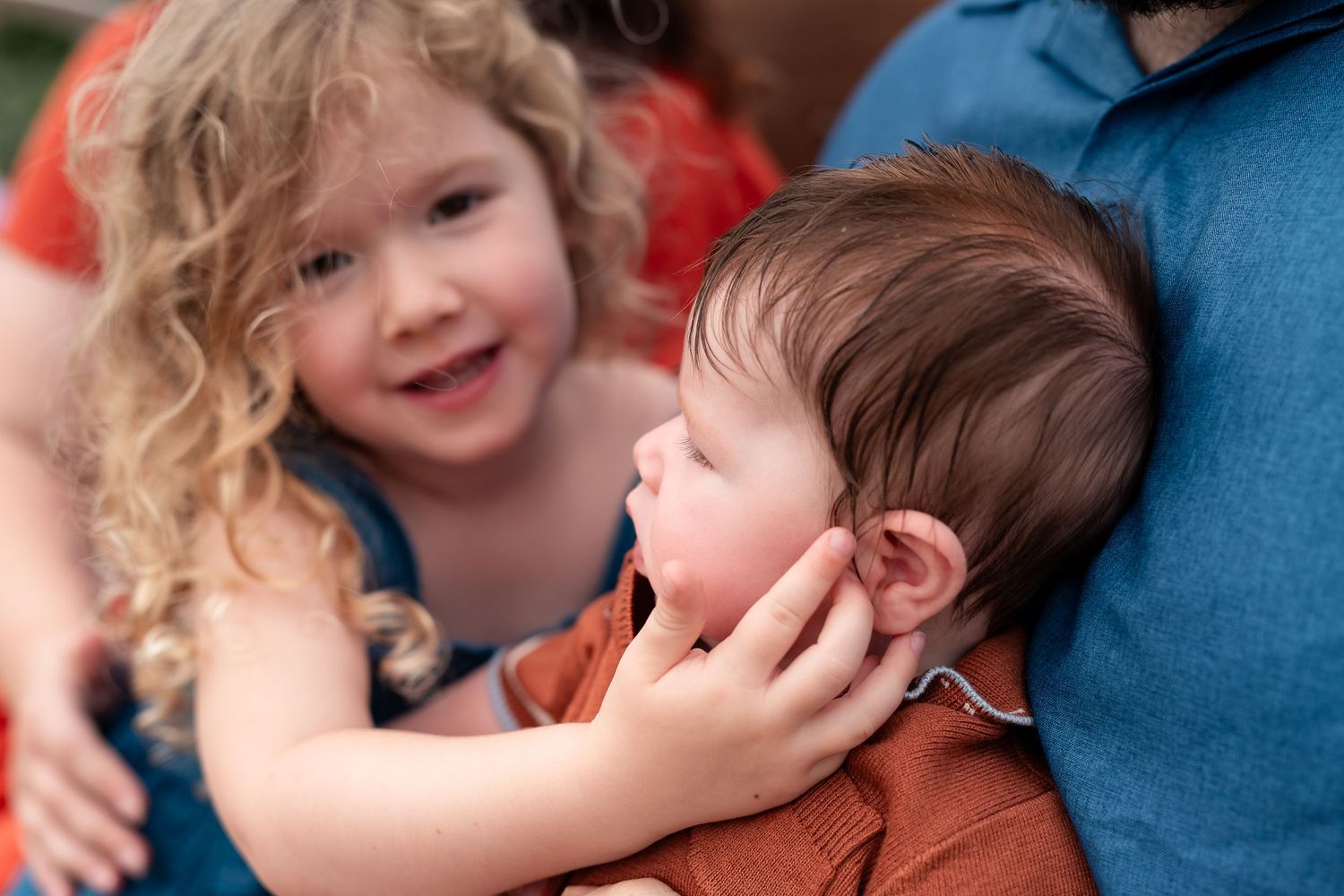 A curly-haired toddler girl lovingly touches the face of a baby being held by an adult in an outdoor setting.