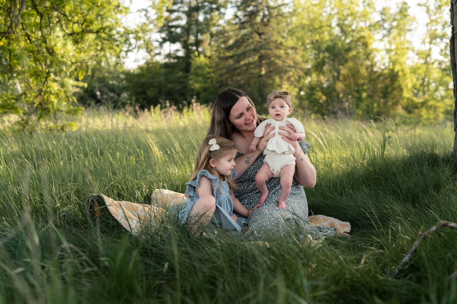 Mother sitting in lush green meadow holding baby while young daughter leans in during golden hour family portrait.