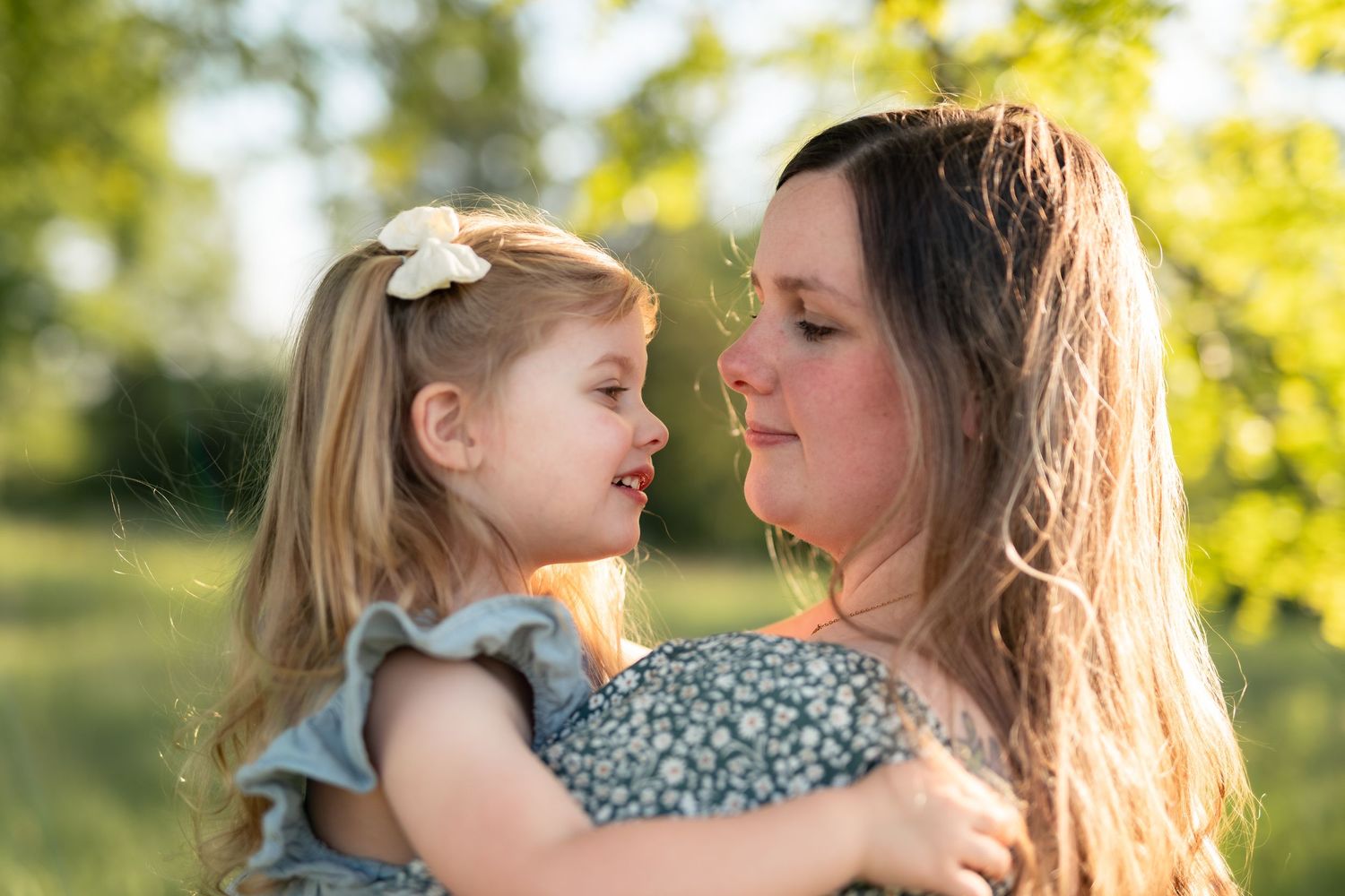 A mother holds her young blonde daughter outdoors in golden sunlight, both smiling at each other in a green park.