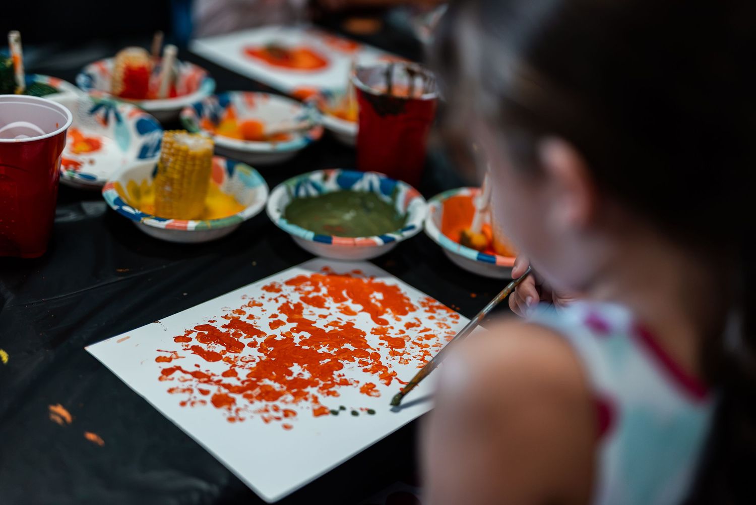 Young girl painting with orange and red colors on white paper at a craft activity table.