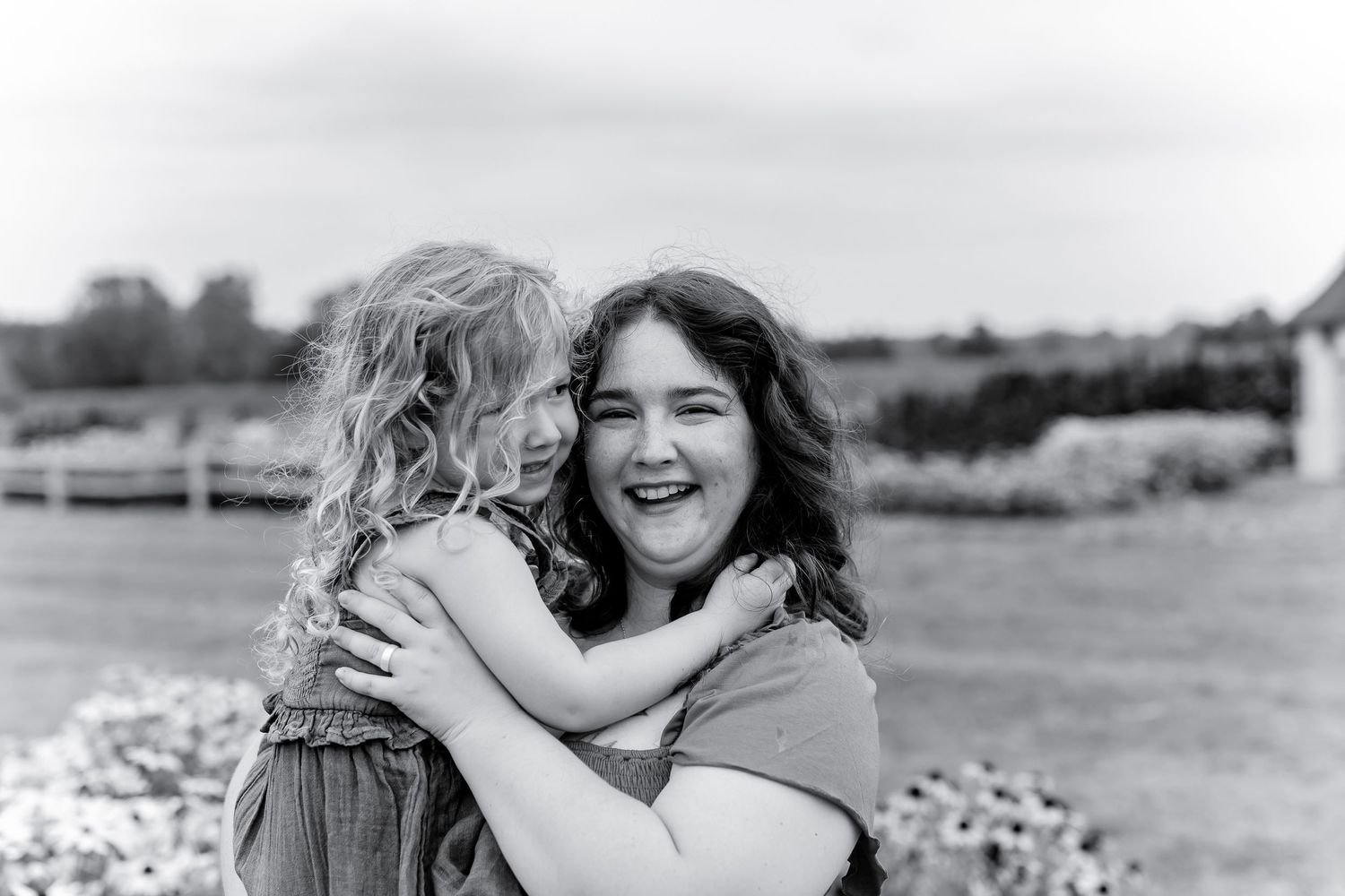 A black and white photo of two girls laughing and hugging outdoors in a garden setting.