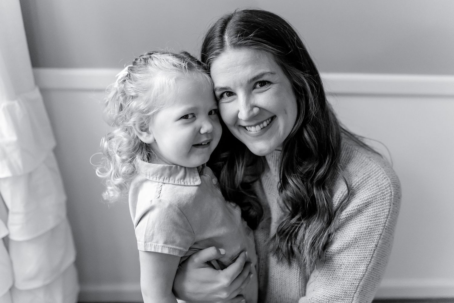 Black and white portrait of a smiling mother hugging her curly-haired toddler daughter indoors.